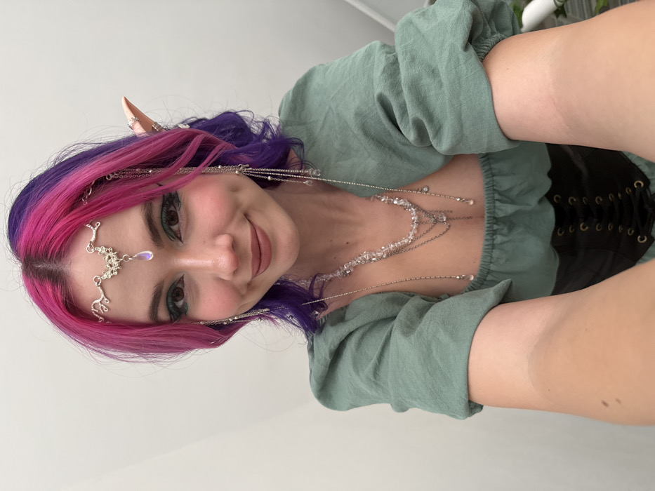 A vibrant selfie captures a person adorned in fantasy-inspired attire, smiling directly at the camera. The image is oriented with the person's head to the left and body extending to the right, suggesting it was taken from a portrait-style perspective and then rotated.
The person has striking two-toned hair, featuring bright fuchsia pink on top that blends into a deep purple underneath and towards the ends. A delicate silver-toned metal headpiece rests on their forehead, characterized by an ornate, possibly vine-like or floral design, from which an iridescent, teardrop-shaped gem hangs centrally. The headpiece also features fine chains that extend back along the sides of their head.
Visible on their ears are pointed prosthetic elf ears, adorned with small silver-toned rings or cuffs, enhancing the fantasy aesthetic. Their face shows a friendly smile, with rosy blush on the cheeks and subtle pink lipstick. Their eyes are accentuated with dark eyeliner and possibly a hint of green or teal eyeshadow on the lower lash line.
Cascading down from the headpiece and around their neck are multiple layers of silver-toned chains and necklaces, many featuring clear, faceted beads or crystals. These chains create a delicate, layered effect over their chest and shoulders.
The person is wearing a light sage green or muted mint green top with voluminous, gathered sleeves, reminiscent of a peasant or renaissance style blouse. Beneath this, a dark, likely black, corset or bodice is visible, tightly laced up the front, further emphasizing the historical or fantasy costume.
The background is a simple, plain white or very light-colored wall, providing a clean contrast that highlights the person and their elaborate ensemble. The overall impression is one of a whimsical, fantasy-themed character.
