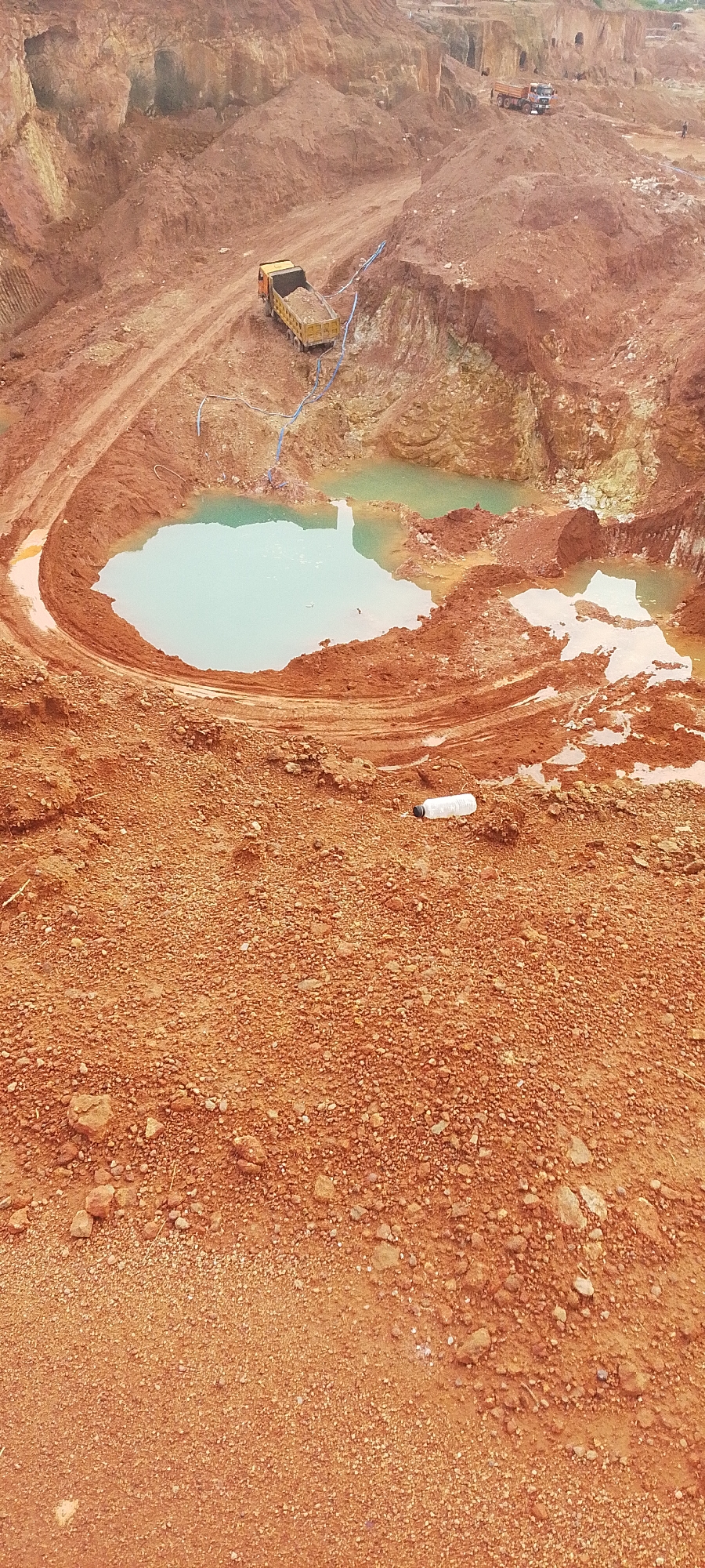 An aerial view depicts a large, open-pit mine with extensive red soil and rock formations. A body of turquoise-colored water has accumulated in a large depression within the mine. Two dump trucks are visible: one is yellow and orange and appears to be partially filled with soil, situated on a dirt path; the other, larger truck is orange and red, positioned on a higher level of the mine. Blue hoses snake across the terrain, likely for water management. In the foreground, a white plastic bottle lies on the dusty ground. The overall scene suggests ongoing excavation and mineral extraction operations.