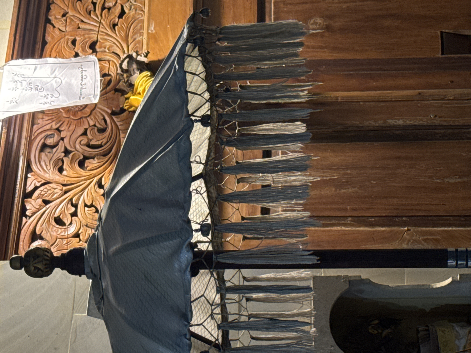 The image shows an umbrella adorned with tassels, positioned in front of ornately carved wooden panels and a white sheet of paper with writing in an unknown language. The handle of the umbrella is visible, suggesting it is standing upright. The wooden panels display a detailed floral or plant-based design. The environment appears to be indoors, with a glimpse of what could be a room or another structure reflected on the lower portion of the image. The location is Kuta Selatan, Indonesia.