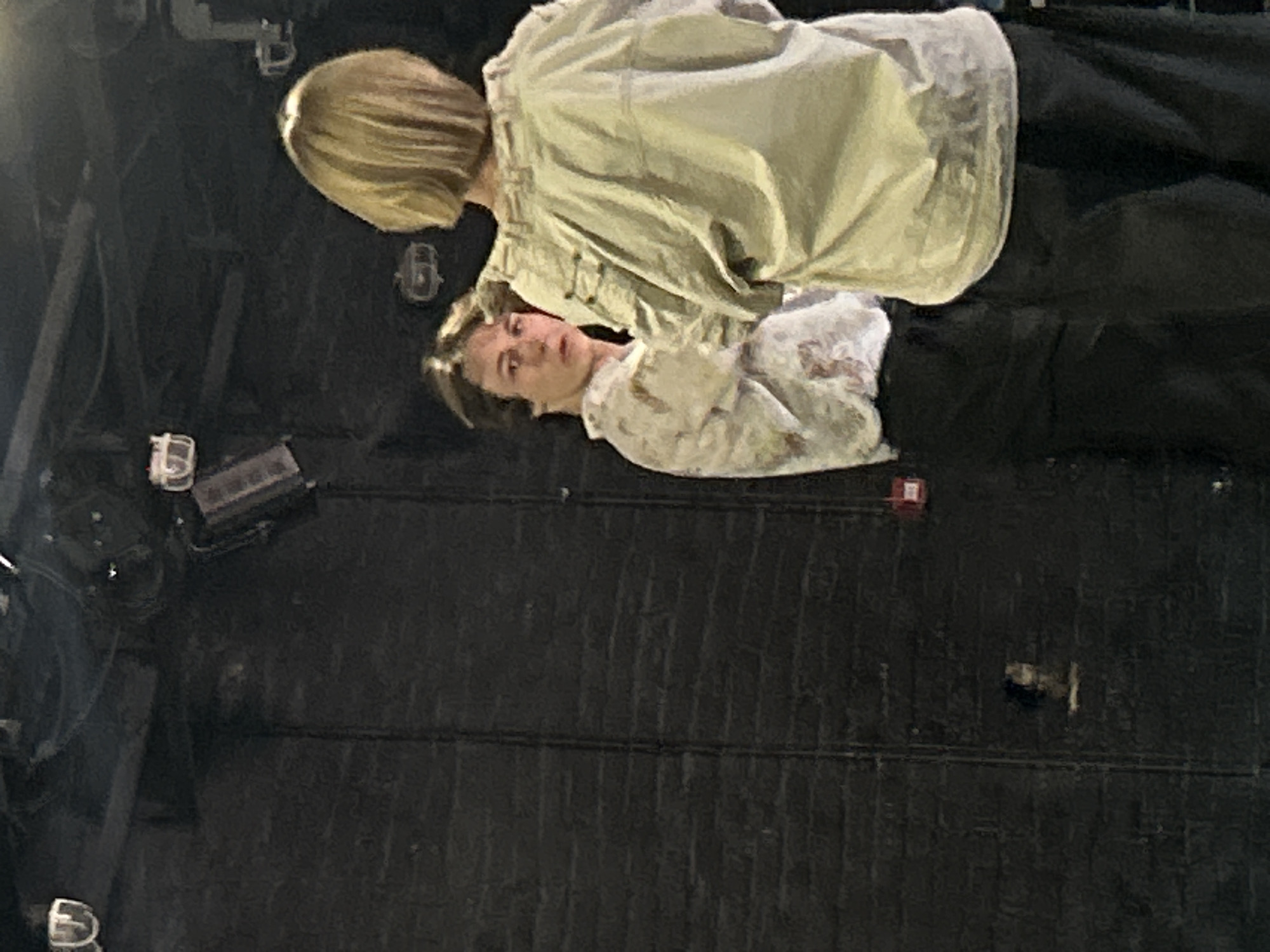 This image shows two people, appearing to be a man and a woman, lying down on a dark surface. The woman, on the bottom, has short brown hair and is looking towards the camera. She is wearing a white shirt with a floral pattern. The man, on top, has blonde hair and is leaning over the woman. He is wearing a light-colored jacket over a dark shirt. Their bodies are positioned close together, suggesting intimacy or a shared pose.
The background is dark and appears to be a textured surface, possibly brickwork or a stage floor. There are some lights and equipment visible in the background, hinting that this might be a performance space, studio, or backstage area. The lighting in the scene is dim, with some light sources creating shadows.
The overall mood of the image is calm and perhaps a little somber, due to the dim lighting and the subjects' relaxed, reclined positions. The individuals' faces are visible, with the woman's expression being neutral and the man's face partially obscured by his posture. There is no visible text in the image. The time of day and weather are not discernible from the image.
