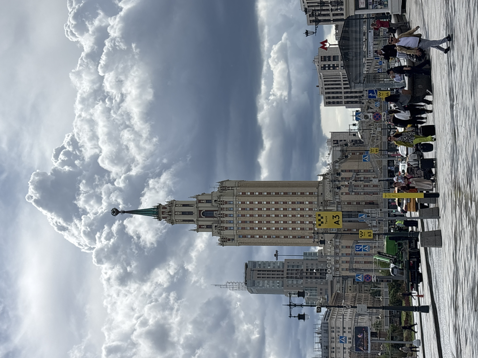 This outdoor shot captures a bustling scene in Moscow, Russia, featuring the Kotelnicheskaya Embankment Building. The foreground shows a wide, paved pedestrian area with various street signs and lamp posts. People are walking and interacting, some with luggage suggesting they may be tourists. The Kotelnicheskaya Embankment Building stands prominently in the mid-ground, with its ornate architectural details. The sky above the building is dramatic, with a mix of dark and puffy white clouds. In the background, other buildings can be seen, indicating the urban setting.