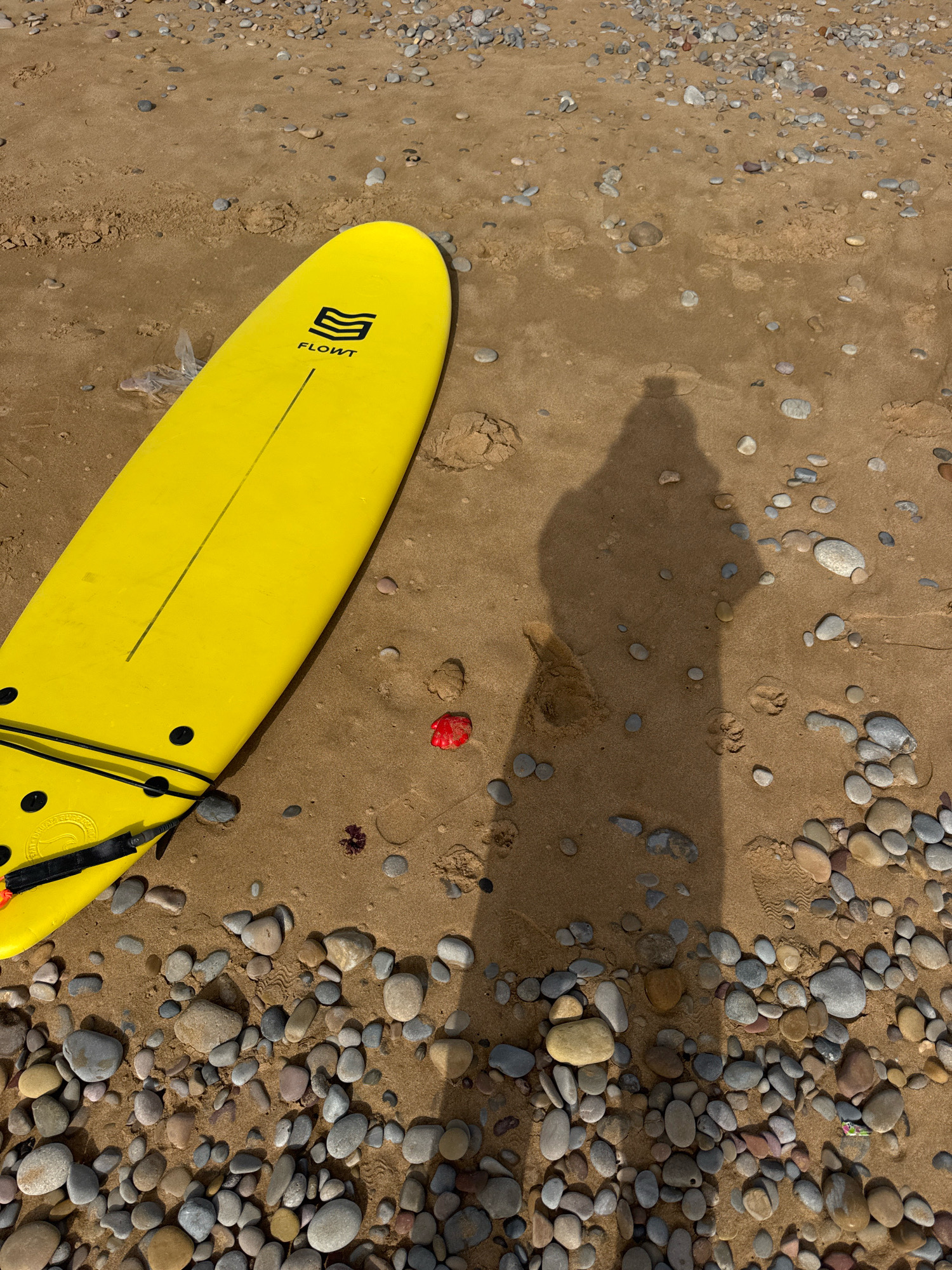 A bright yellow surfboard lies diagonally on a sandy beach in Tamri, Morocco. The surfboard has a black logo and a single black line down its length. Its tail is visible at the bottom left, with black fins and a leash attached. In the foreground, a dense collection of smooth, grey and brown pebbles covers the sand. Further up the beach, the sand is less populated with stones and shows ripples from the tide. A red object, possibly a piece of shell or seaweed, is visible in the sand near the center of the image. A faint shadow of a person, likely the photographer, stretches across the sand from right to left, indicating an overhead light source, possibly the sun. The scene is bathed in natural light.