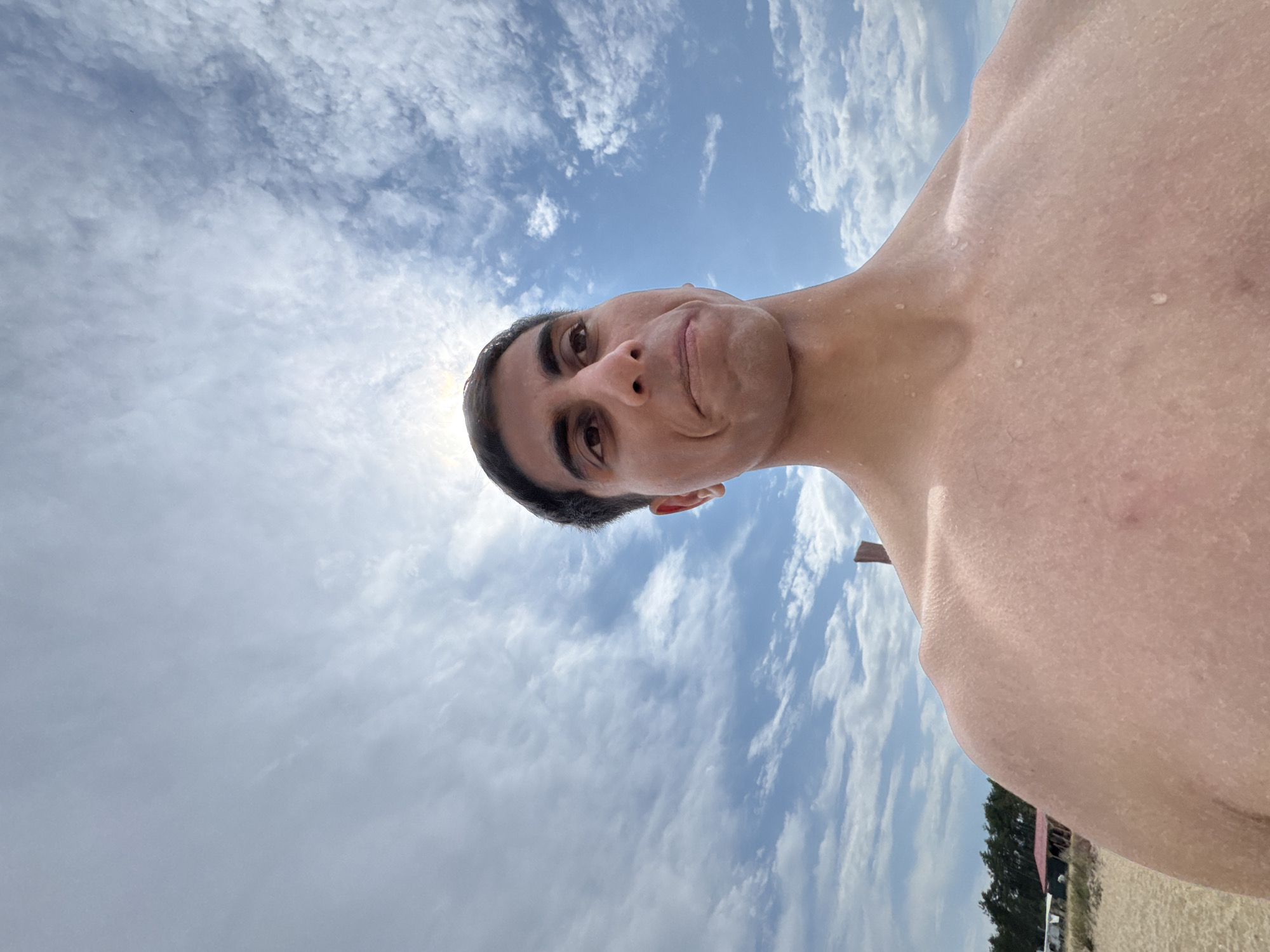 A close-up, low-angle, wide-lens selfie shows a shirtless man with wet skin, facing the camera against a bright, cloudy sky. He has dark hair and is looking slightly off to the right with a neutral expression. The background reveals a beach with sand, trees, and a small structure with a red roof. The scene is consistent with a location in Sozopol, Bulgaria, likely on the coast, given the sandy beach and clear skies. The man's body is well-defined, and water droplets are visible on his skin, suggesting he may have been swimming. The sun is visible behind his head, creating a slight glare.