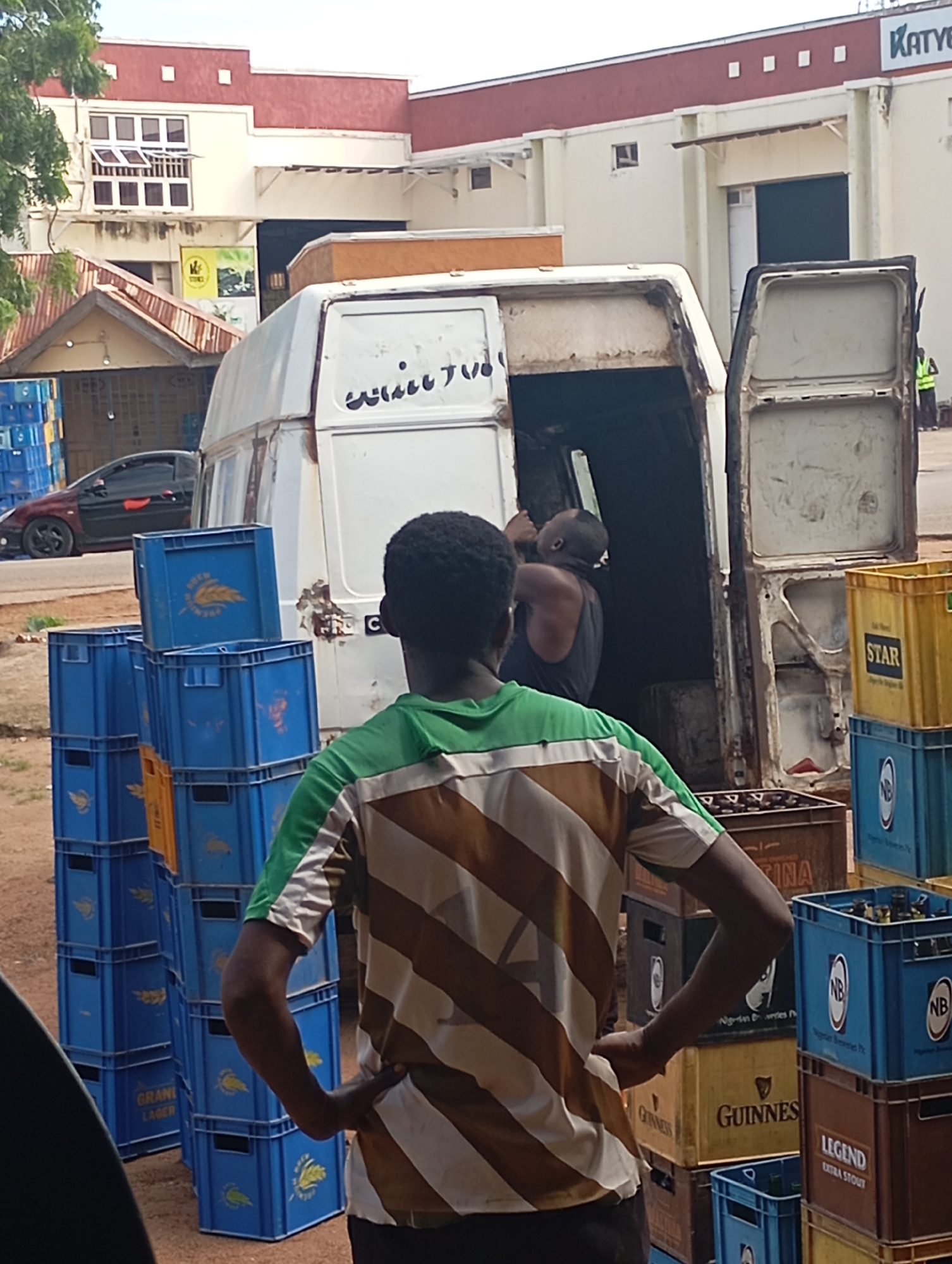 In Shendam, Nigeria, a man wearing a green and brown striped shirt stands with his back to the camera, facing a white delivery van with its doors open. The van is laden with crates, some of which appear to be beer crates. To the left of the van, a stack of blue crates is visible, with a black car parked behind them. In the background, a building with a red and cream facade stands, featuring several windows and an awning. Another man, wearing a dark tank top, is partially visible inside the van, appearing to be involved in the unloading or loading process. The ground is dusty and uneven, suggesting an outdoor environment, likely a commercial area or storage facility.