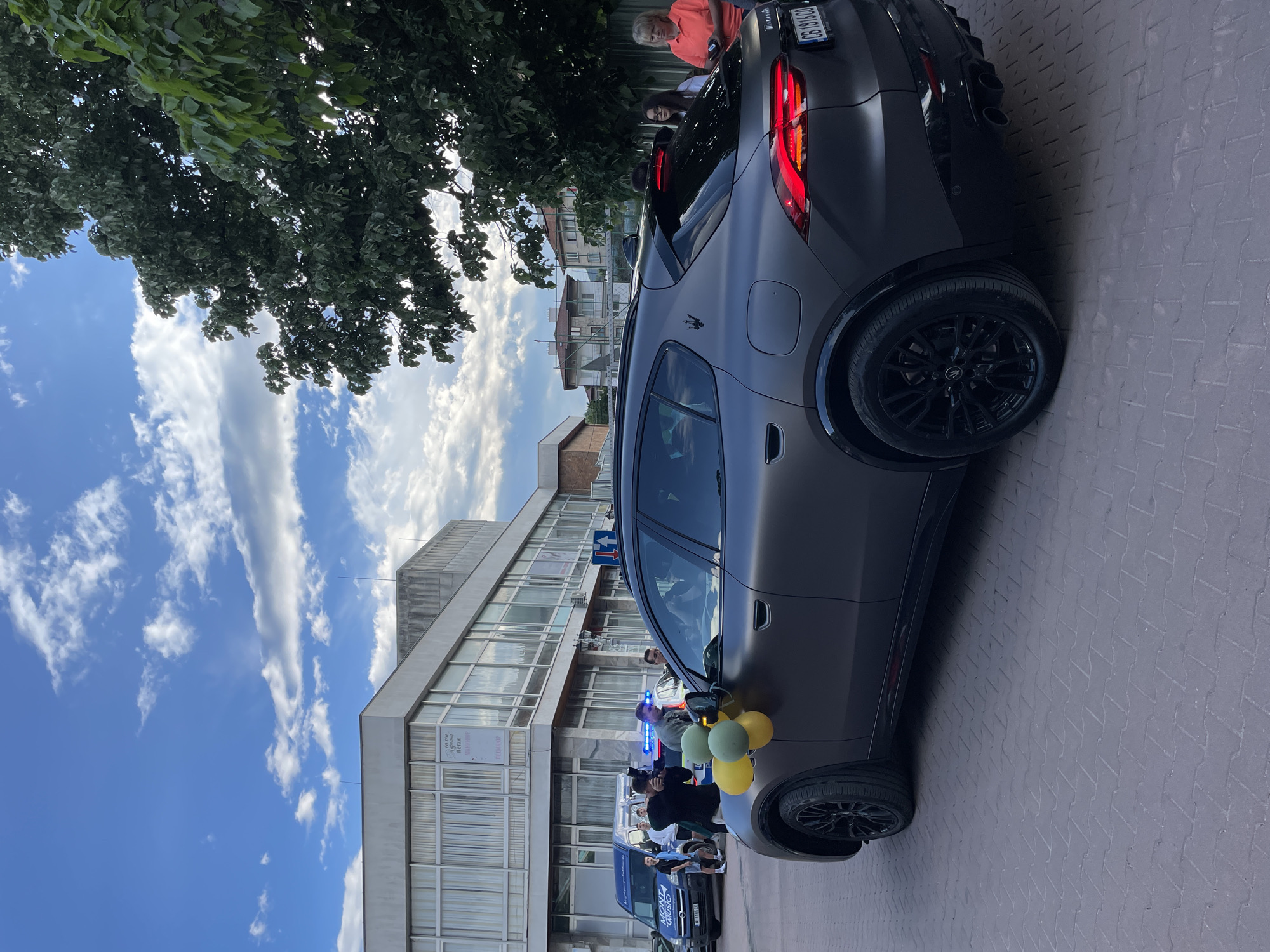 The image shows a dark gray Maserati Grecale parked on a brick-paved area in Zlatograd, Bulgaria. The vehicle is positioned adjacent to a building featuring large windows and what appears to be posters or advertisements. A small group of people stands around the car. Balloons are attached to the vehicle. A blue van with branding on the side is parked nearby. Above the building, the sky is visible with scattered clouds. A tree partially obscures the background on the left side of the image. The license plate number of the Maserati Grecale is CB 1816 CB.