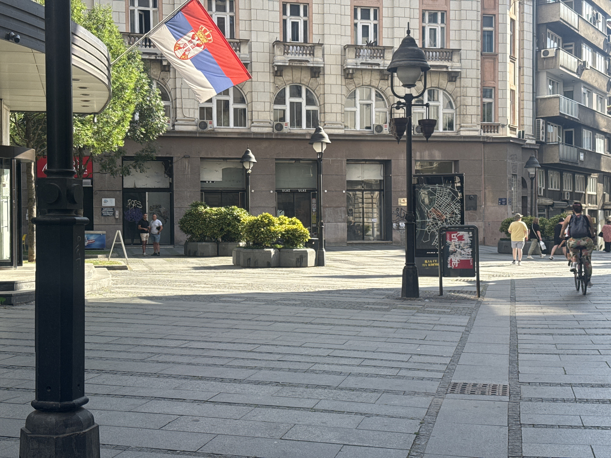 The image depicts a street scene in Belgrade, Serbia, specifically Knez Mihailova Street, a well-known pedestrian promenade. In the foreground, a black lamppost stands on the left side of the frame, with a sidewalk made of large grey stone slabs. A Serbian flag, featuring the national colors of red, blue, and white with the coat of arms, is prominently displayed, attached to a building.
Behind the lamppost, a modern building with large windows and balconies is visible. Across the street, other buildings exhibit varied architectural styles, some with ornate facades and others with more contemporary designs. Several street lamps and planters with green foliage are also present.
Several individuals are walking on the street. On the right side of the image, a person is riding a bicycle, facing away from the camera. Further in the background, other pedestrians are seen walking in various directions. The street is well-lit by sunlight, casting shadows across the stone pavement, indicating it is daytime. The overall impression is of a bustling urban environment, characteristic of a popular city center.