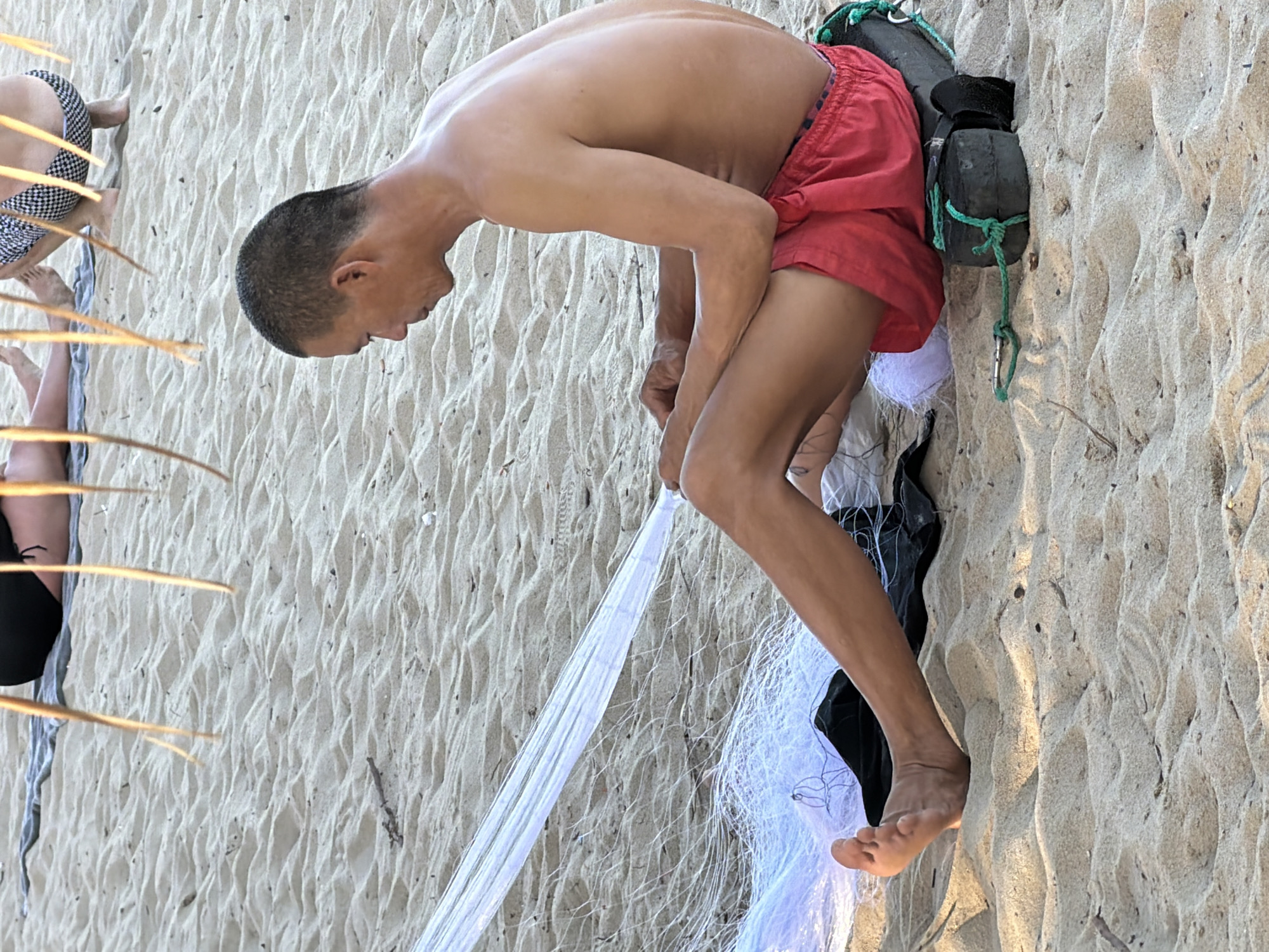 An adult male is sitting on a sandy beach in Nha Trang, Vietnam, working with a white fishing net. He is wearing red shorts and is shirtless. His legs are extended, and he is using his hands to manipulate the net. The net is spread out in front of him and draped over some dark material and a black, cylindrical object with green rope tied to it. In the background, out of focus, are other beachgoers, including one person wearing a checkered swimsuit and another wearing dark bottoms. The sand is textured with small waves and ripples, and some dried vegetation is scattered on the ground. The lighting suggests it is daytime.