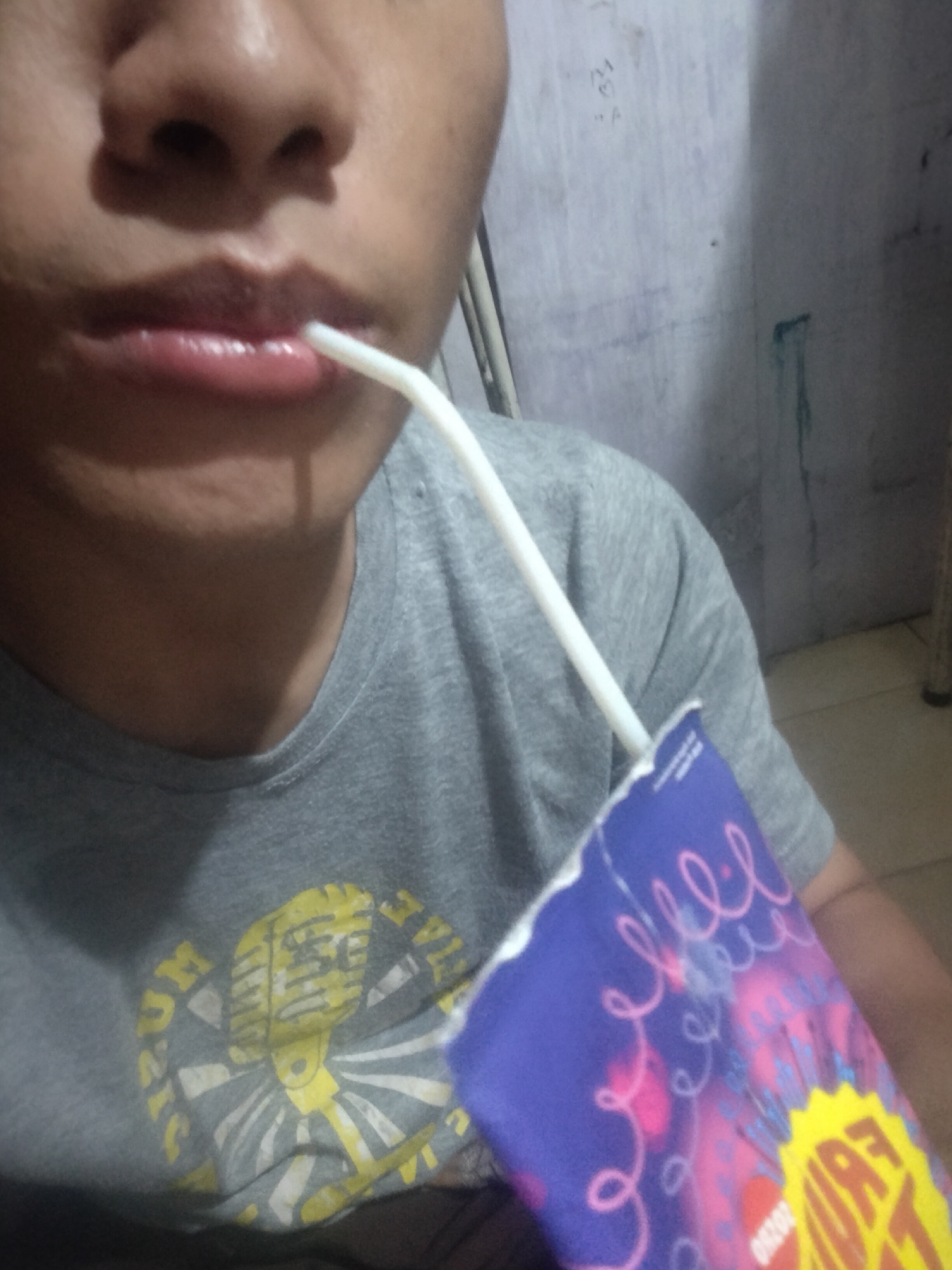The image is a close-up, waist-up shot of a person wearing a gray t-shirt. The t-shirt has a graphic of a yellow microphone surrounded by radiating lines and text, partially obscured by a blue and purple box held in the person's hand. The person is using a white straw to drink from the box, which has colorful swirling patterns and text. The background is a wall with some blue markings and a dark greenish stain. The lighting is dim, suggesting an indoor setting, possibly a residential area in the city of Medan, Indonesia, based on the provided hints. The focus is on the person's interaction with the drink.