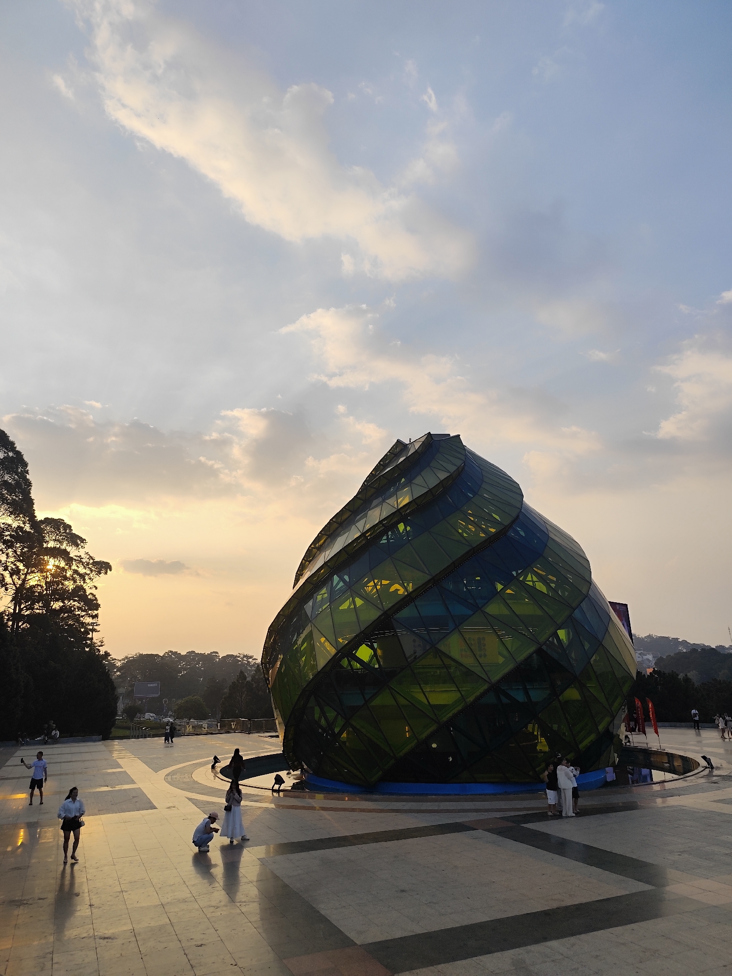 The image displays Lam Vien Square in Da Lat, Vietnam, during sunset. A large, distinctive, spiraling glass building, predominantly green and blue, and known as the "Artichoke Bud," occupies the central-right portion of a wide, light-colored tiled plaza. The sky above features scattered clouds, with warm golden and orange hues near the horizon transitioning to lighter blue. Approximately twelve individuals are observed on the plaza; some are walking, others are standing or kneeling, appearing to take photographs. One individual in a white t-shirt is kneeling in the mid-ground. To the left, tall evergreen trees are silhouetted against the setting sun. In the background, forested hills and scattered distant buildings are visible. The low sun casts warm, golden light across the scene, creating reflections on the tiled ground and the glass structure.