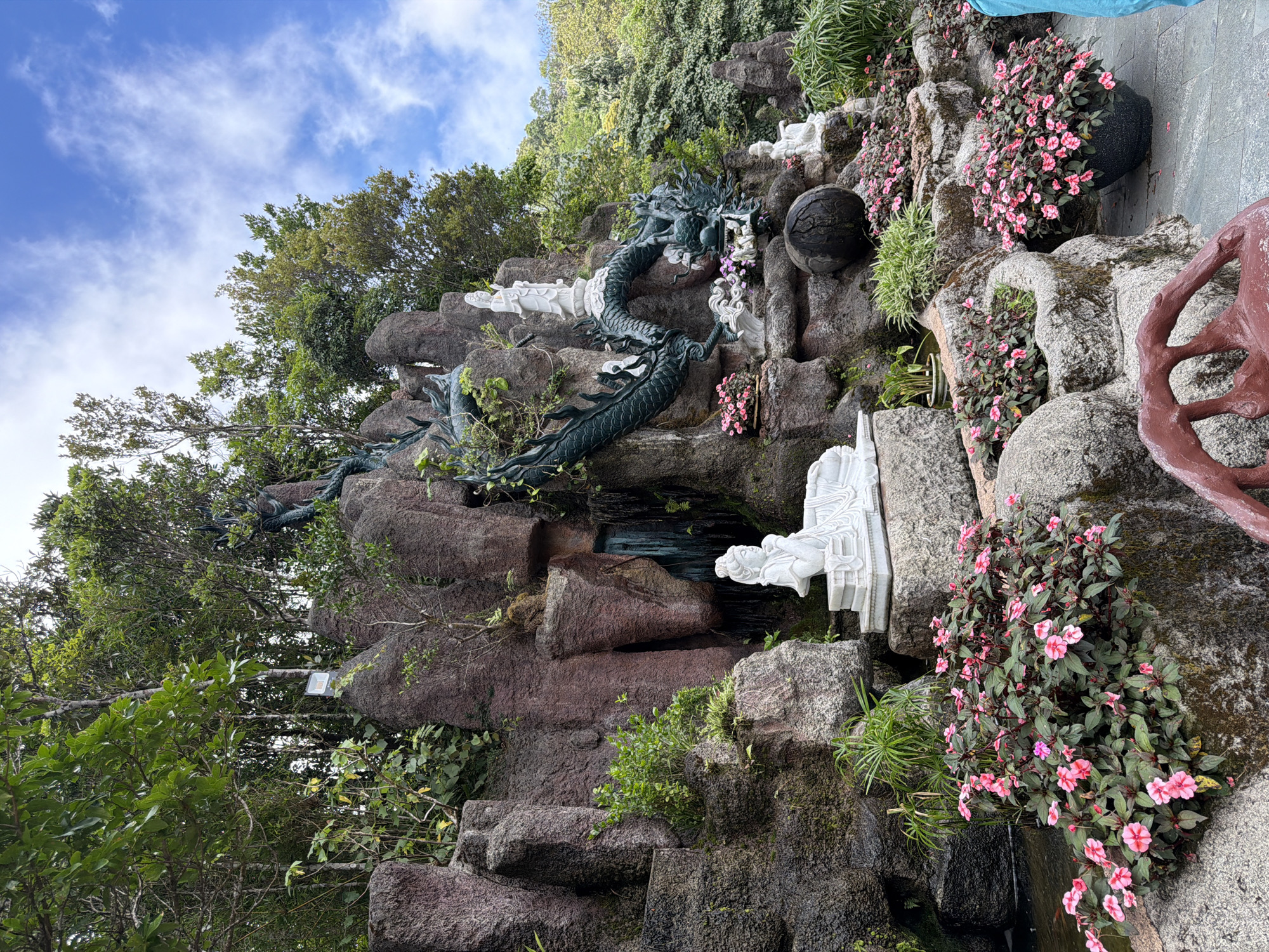 The image depicts a scenic outdoor area, likely a garden or temple complex, featuring a prominent rock formation that resembles a waterfall. A large, dark green, serpentine dragon sculpture is coiled around the rock formation, with its head emerging near the top. Beside the dragon, on a raised platform, stands a white marble statue of a female figure. Below the dragon, on another level of the rock formation, lies a reclining white marble statue of a woman. Several smaller white marble figurines are also visible among the rocks and plants. The foreground is adorned with numerous pots of pink impatiens flowers, and a dark, spherical object is situated on the rocks. The background is lush with green trees and foliage, against a bright blue sky with scattered white clouds. The location is identified as a Buddhist temple or similar spiritual site in Da Nang, Vietnam.