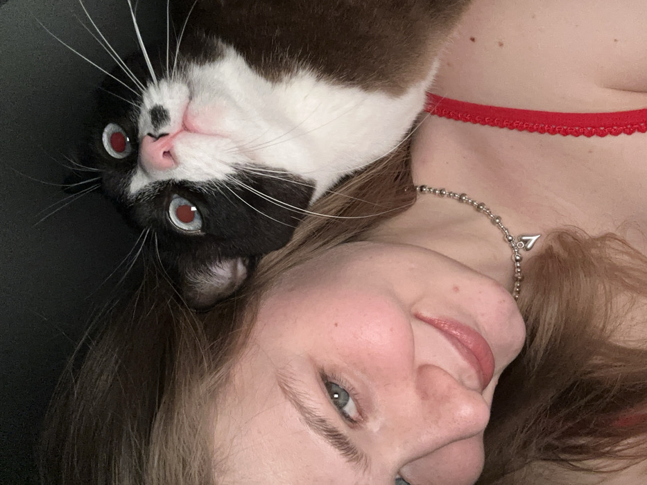 A close-up, selfie-style photograph depicts a young adult female and a domestic cat, likely taken indoors in low light conditions as indicated by the cat's red-eye effect. The scene is located in Noviy, Russia.
The female subject occupies the left side of the frame, angled towards the viewer with a slight smile. She has light-toned skin, light-colored hair, and blue-green eyes. She wears a silver-toned beaded necklace with a small heart pendant. A red garment with a scalloped edge is visible on her right shoulder.
The domestic cat, a bicolor (black and white) animal, is positioned on the right side of the frame, partially obscured by the female's hair and head. Its head is tilted upwards, looking towards the camera with dilated pupils exhibiting a pronounced red-eye effect. The cat has prominent white whiskers and white fur on its muzzle, chin, and chest. The background is uniformly dark and featureless. The subjects are in close proximity, suggesting an interaction.