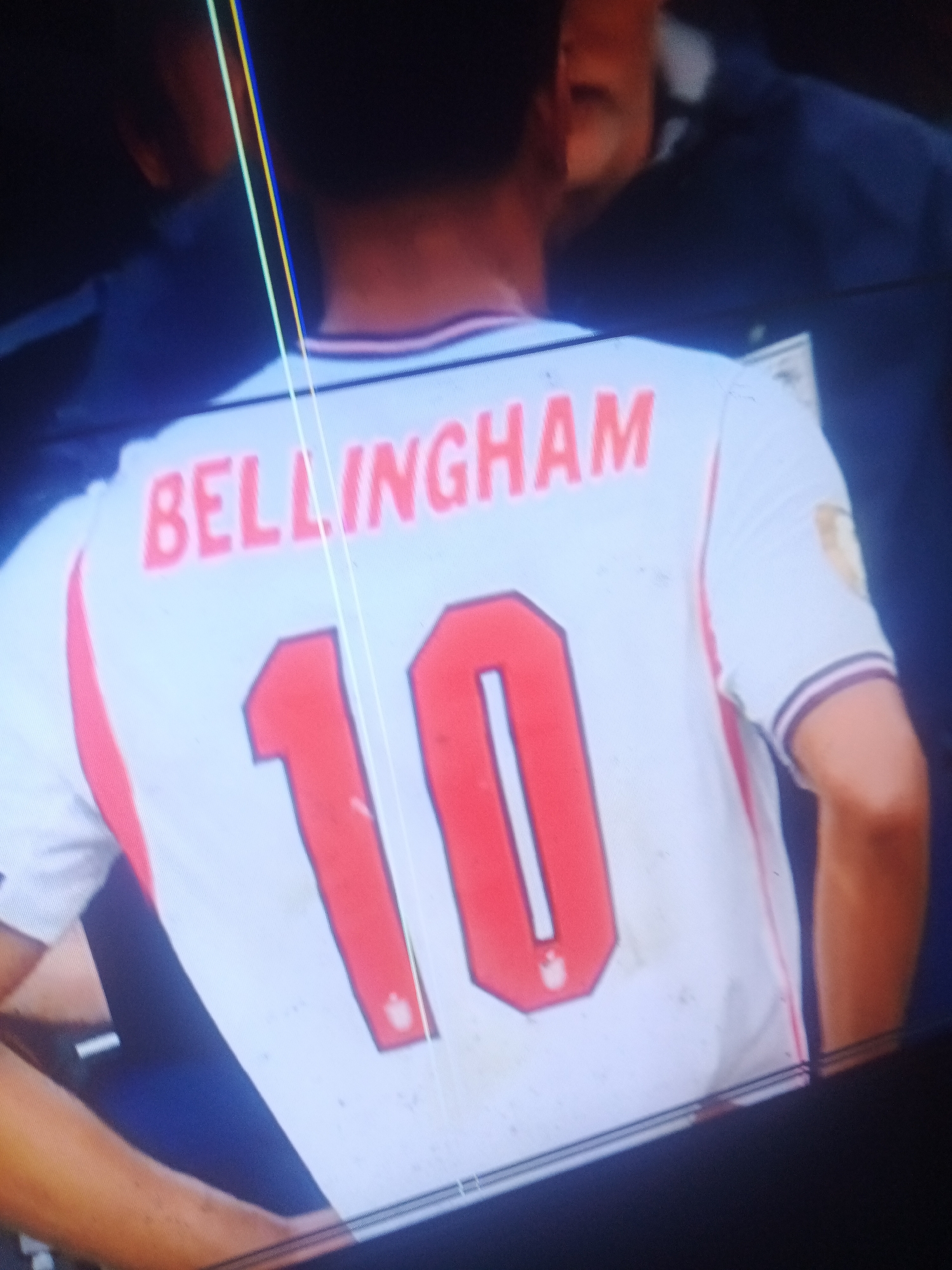 The image shows a close-up of the back of a white soccer jersey with red trim and lettering. The name "BELLINGHAM" is visible in red capital letters across the upper back, with the number "10" prominently displayed below it in large red numerals with a white outline. A golden emblem or patch is visible on the upper right shoulder of the jersey. The fabric of the jersey appears slightly worn or dirty in places.
The setting is unclear, but the image quality suggests it might be a screenshot from a video or a photograph taken from a distance, possibly on a screen. The background is dark and out of focus, with hints of other people or figures present. The lighting is somewhat dim. There are also some thin, colored vertical lines overlaid on the image, likely artifacts from the recording or display method.
The context provided states the location as Jalingo, Nigeria. Without further visual cues in the image itself, it's impossible to confirm or deny this location based solely on the provided media. No specific time of day or weather conditions are discernible. The activity is likely a sporting event, specifically a soccer match, given the jersey. No emotions are directly conveyed by the visible subjects.