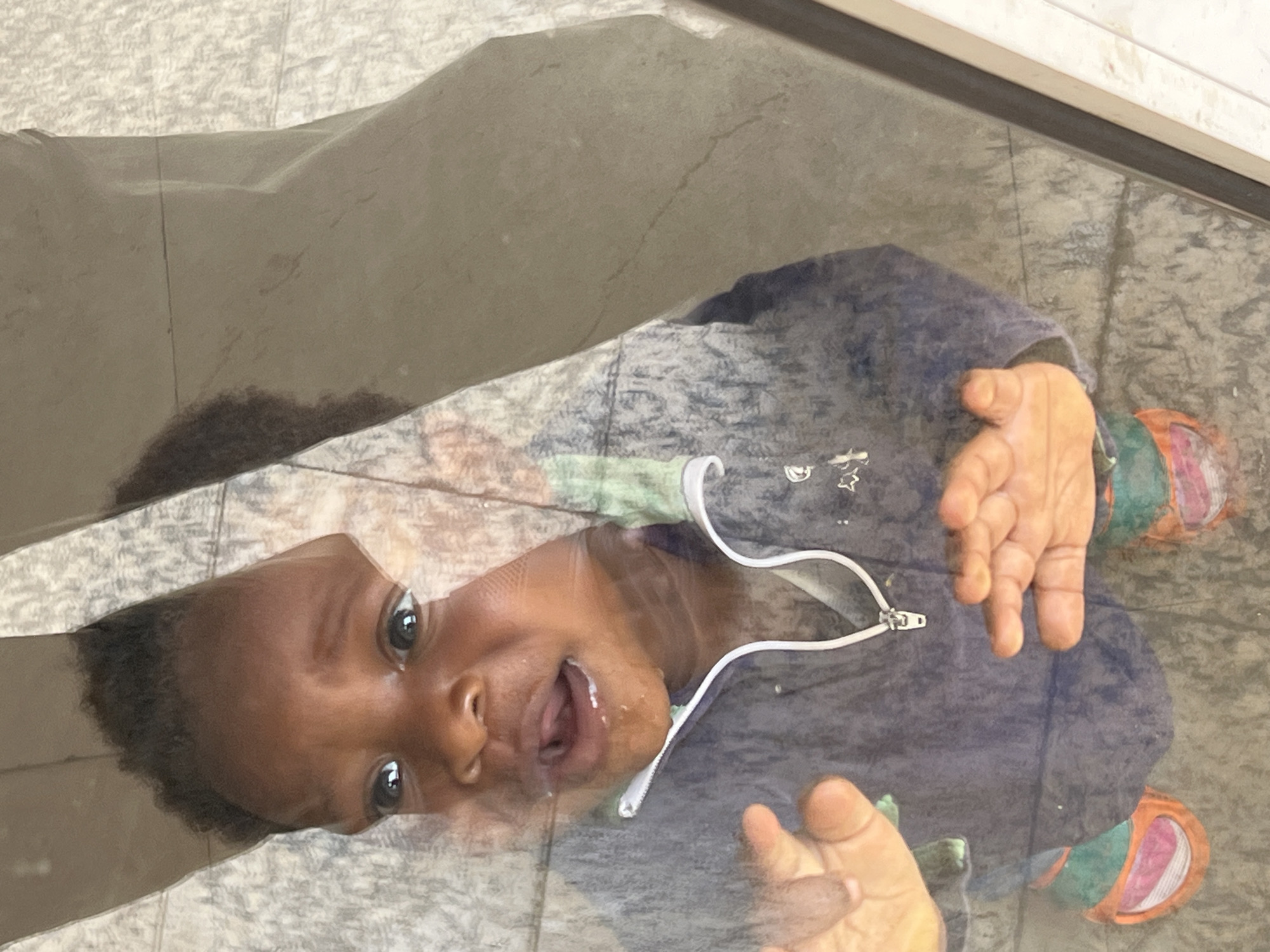The image captures a young child, likely a toddler, looking upwards directly at the camera through a clear, reflective surface, possibly glass or plastic. The child has dark skin, short dark curly hair, and wide, curious eyes. Their mouth is open, revealing teeth and tongue, with a noticeable streak of saliva or drool around the mouth.
The child is wearing a dark blue or grey hooded jacket featuring a white zipper down the front and white drawstrings on the hood. Small, light-colored patterns, possibly stars, are visible on the left chest area of the jacket. The child also appears to be wearing colorful trousers or leggings, with visible sections of green and orange/pink fabric. Both of the child's hands are open and raised, positioned on either side of their head, in a gesture that suggests reaching out or playful interaction.
The background, seen through and around the child and the clear surface, appears to be a light-colored, textured floor or ground, with some darker patches and faint lines, possibly indicating tile or concrete. The lighting in the image suggests it is daytime. The overall scene conveys a moment of innocent curiosity and engagement from the child. The provided location context is Gura Top, Nigeria. There is no visible text in the image.