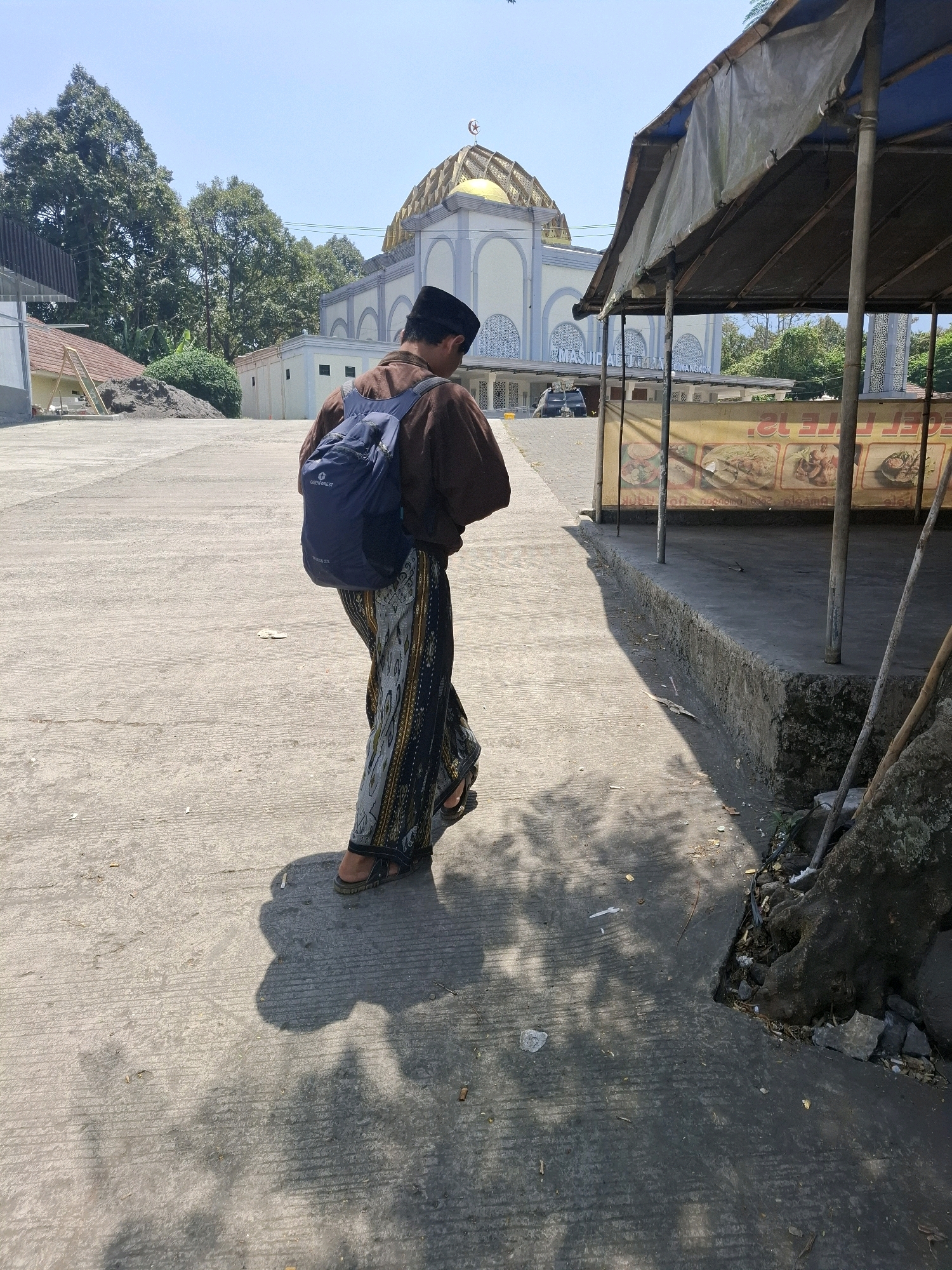 A person wearing a brown shirt, black peci (cap), and a patterned sarong walks away from the camera on a paved walkway. The person is carrying a blue backpack and wearing sandals. In the background, a large mosque with a golden dome and white walls is visible. The mosque has text that reads "MASJID AL-FATIH..." above its entrance. To the right of the walkway, there is a covered stall with a yellow banner displaying images of food. Trees and a clear blue sky are also present in the scene. The overall environment appears to be an outdoor public space, possibly near a place of worship or a market. The location is identified as Sukalarang, Indonesia.