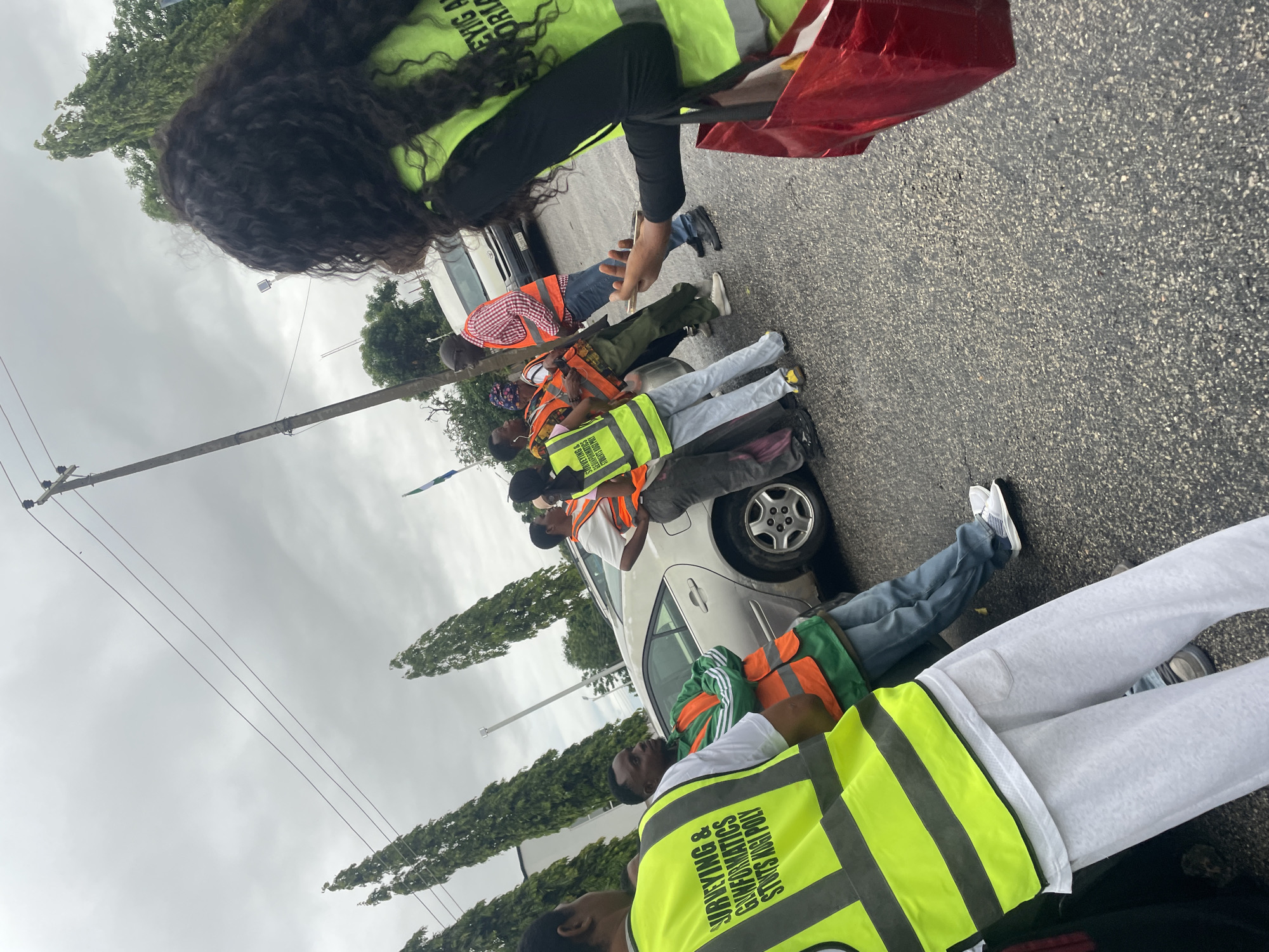 In Lokoja, Nigeria, a group of people are gathered outdoors on an asphalt surface, possibly a road or parking lot. Several individuals are wearing bright yellow safety vests with reflective stripes. One person in the foreground, facing away from the camera, wears a yellow vest over a white t-shirt and light gray sweatpants. Another person, partially visible in the foreground left, also wears a yellow vest with text that includes "SURVEYING & GEOMATICS" and "STUDENTS NGI. POLY". To the right, a woman with long, dark curly hair, also wearing a yellow safety vest, holds a red shopping bag. Several other people in the middle ground are wearing orange safety vests and appear to be interacting. Behind the group, a silver sedan is parked, and a white van is visible further back. Tall, slender trees line the background, and utility poles with wires are present against an overcast sky.