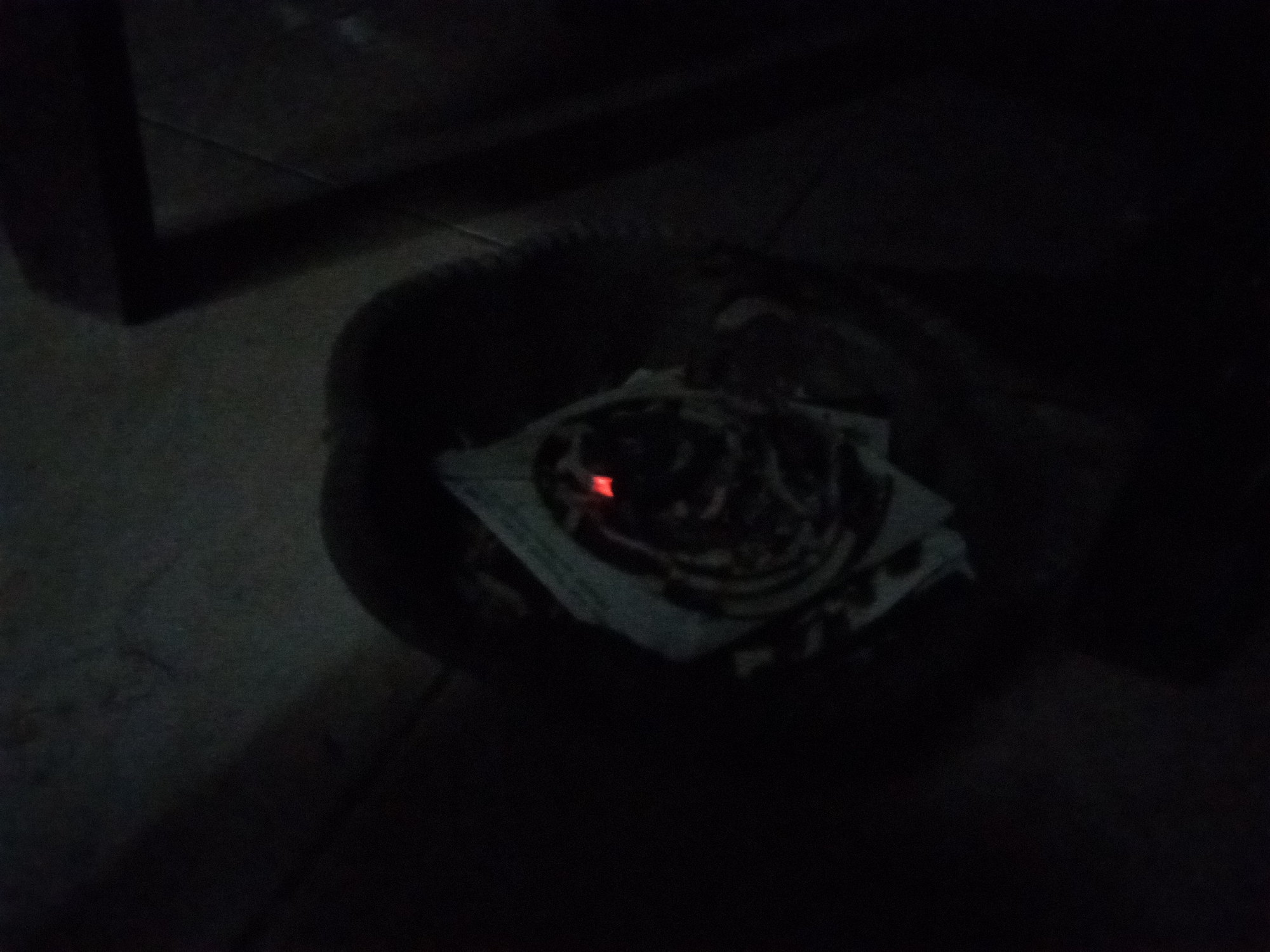 The image depicts a dark environment with a circular object in the foreground, possibly a bowl or a dish, containing a pile of white paper and a small, glowing red ember. The scene is dimly lit, and the background is obscured by shadow, suggesting an indoor setting at night or a poorly lit area. There are no discernible people or specific landmarks that would indicate a location in Pemalang, Indonesia. The composition is focused on the object in the foreground, with the ember being the only source of bright light.