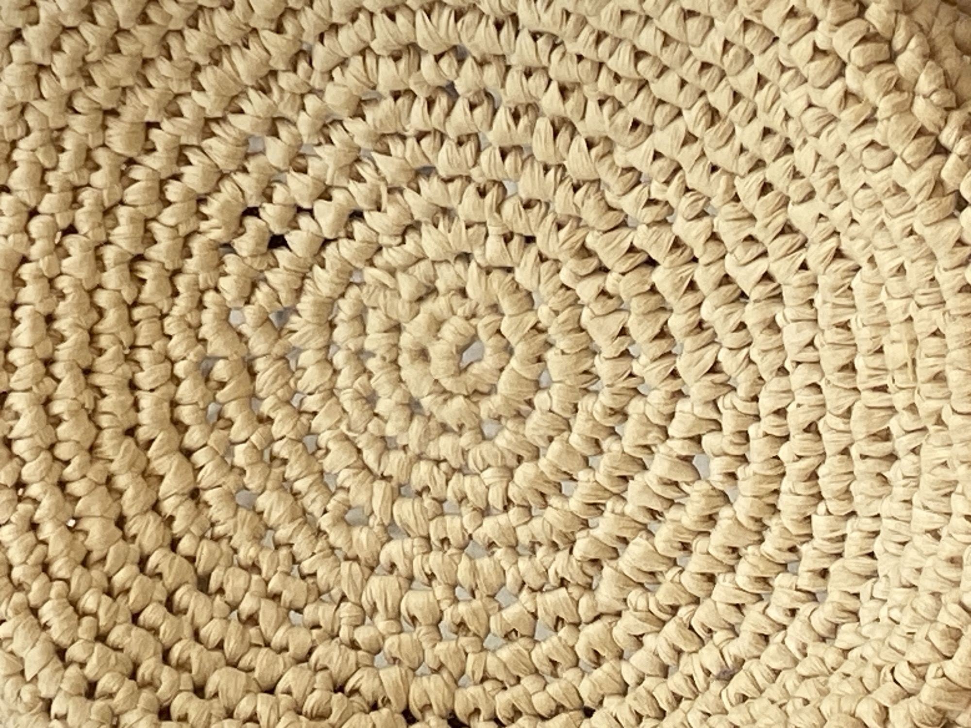 This close-up image displays the intricate texture and pattern of a tightly woven material, likely made from natural fibers such as straw or raffia. The material exhibits a uniform light beige or tan color. The weave forms a distinct spiral pattern, originating from a central point and expanding outwards in concentric, circular rows. Each segment of the weave appears as a small, folded, or bunched piece of fiber, creating a rich, tactile surface with subtle variations in light and shadow. The meticulous arrangement of these individual elements highlights the handcrafted nature of the item. There are no other subjects, objects, or activities visible, as the photograph focuses exclusively on the detail and structure of the woven texture. The lighting is soft and even, accentuating the material's depth and form. Given the context of Alanya, Turkey, this material could be part of a local craft item such as a traditional mat, basket, or a sun hat. No text is present in the image.