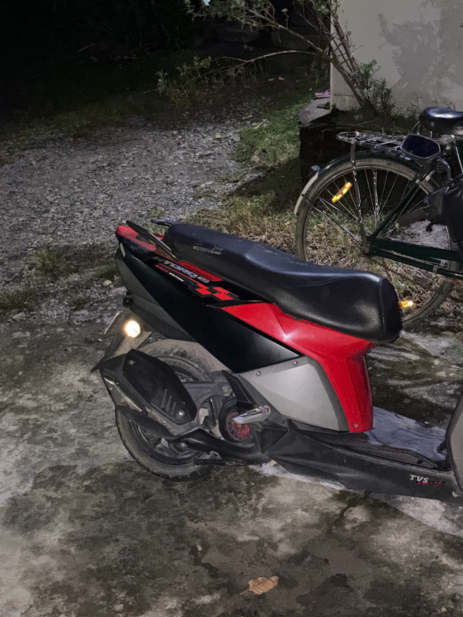 An outdoor, ground-level, nighttime photograph shows a black and red scooter parked next to a dark green bicycle. The scooter is positioned in the foreground, angled slightly to the left. It has a prominent black seat with a red and black patterned detail on the side panel. The rear fender of the scooter is visible, as is the exhaust pipe. The bicycle is in the background, to the right of the scooter, with its rear wheel clearly visible. The bicycle has a black seat and a metal rack over the rear wheel. The ground is a mix of gravel and a damp, concrete-like surface. Vegetation, including grass and some shrubs, is visible in the background and around the vehicles. The lighting suggests artificial light sources are present, casting shadows. The image appears to be taken in Sherfanguri, India.
