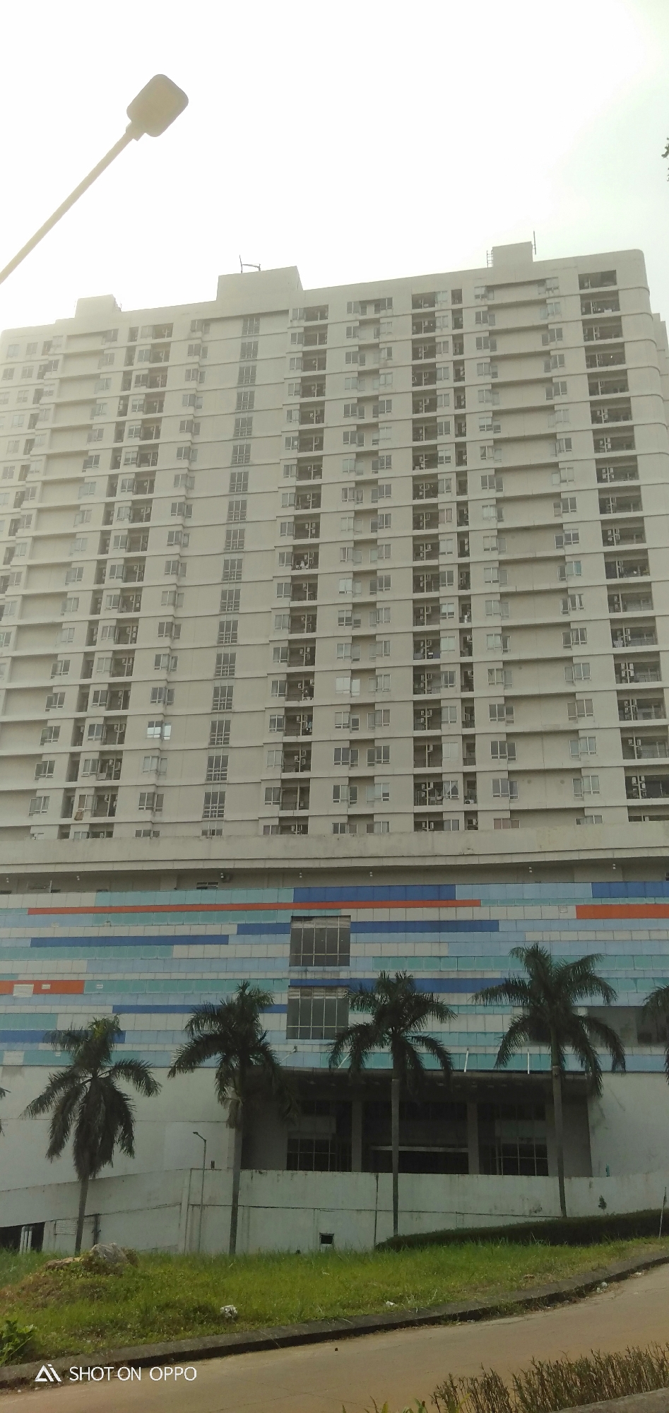 The image shows a very tall, light-colored apartment building in Depok, Indonesia. The building has many windows and balconies, and it appears to be a modern residential complex. In front of the building, there are several palm trees and a colorful facade with blue, white, and orange tiles. The sky is overcast and bright. The image is taken from a low angle, looking up at the building. There are no people visible in the image.