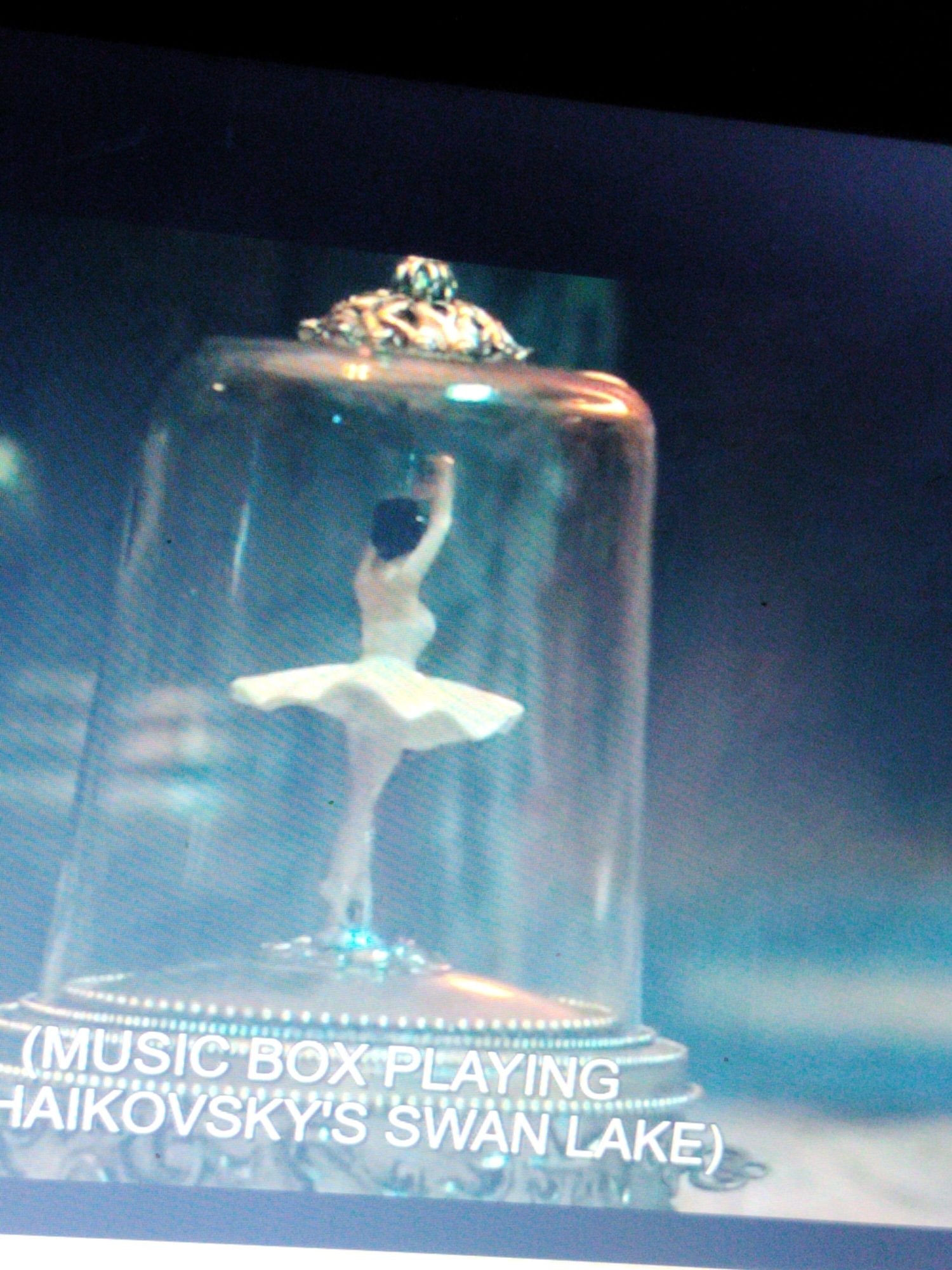 The image shows a close-up of a music box that appears to be a decorative item. Inside a glass dome, a small figurine of a ballerina in a white tutu is posed in mid-dance. The base of the music box is ornate, with what looks like a metallic finish and decorative etching. Text is visible on the base, reading "(MUSIC BOX PLAYING TCHAIKOVSKY'S SWAN LAKE)". The background is dark and blurred, making it difficult to discern any environmental details or the location within Du, Nigeria. There are no people or interactions visible in the image, only the music box and its contents.