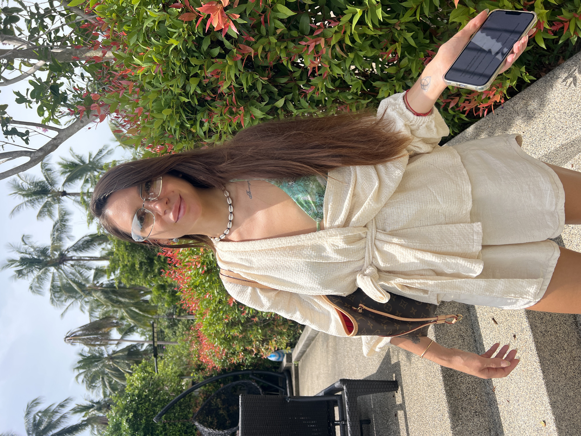 A woman with long brown hair and sunglasses stands in an outdoor setting, possibly a resort or garden, with lush greenery and palm trees in the background. She is wearing a light-colored, wrap-around top over a patterned bikini top, and matching shorts. A Louis Vuitton tote bag is slung over her shoulder, and she is holding a smartphone in her right hand. She has a seashell necklace and several pieces of jewelry on her arms, including a bracelet and a red string tied around her left wrist. The environment suggests a warm, tropical location, consistent with the city of Ban Bang Rak, Thailand. The ground appears to be a tiled pathway with some steps.
