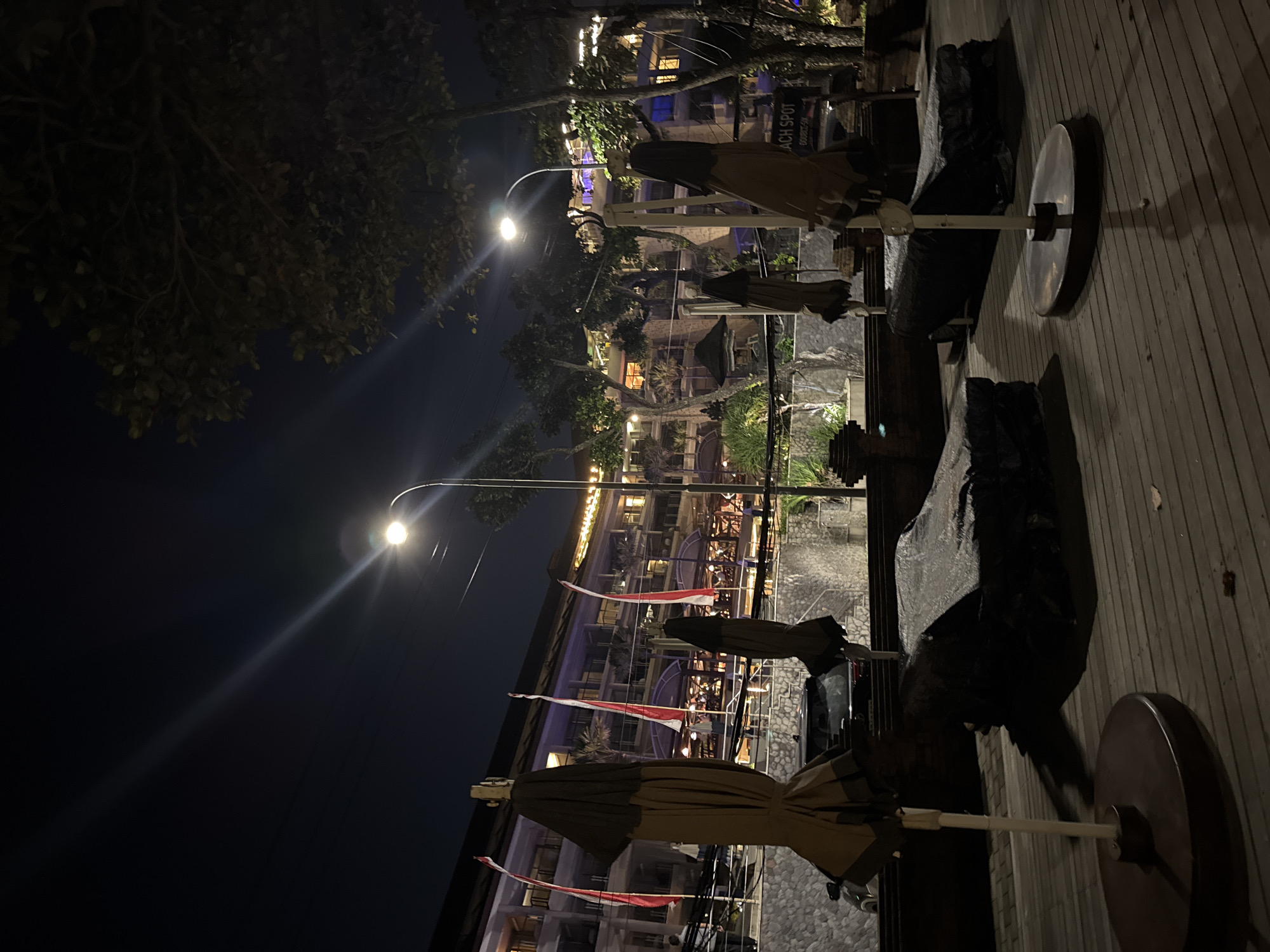 This is a nighttime photograph of a beachside resort in Kuta, Indonesia. The scene is illuminated by streetlights and the lights of the resort building, which is visible in the background. In the foreground, several beach chairs are covered with dark tarps, and closed umbrellas are propped up on stands. The ground is a wooden deck. A stone wall with a tiered structure lines the area in front of the resort. The resort itself appears to be multi-story with balconies and windows lit from within. Red and white flags are displayed along the building's facade. A car is parked near the wall. The overall impression is of a quiet, closed resort at night.