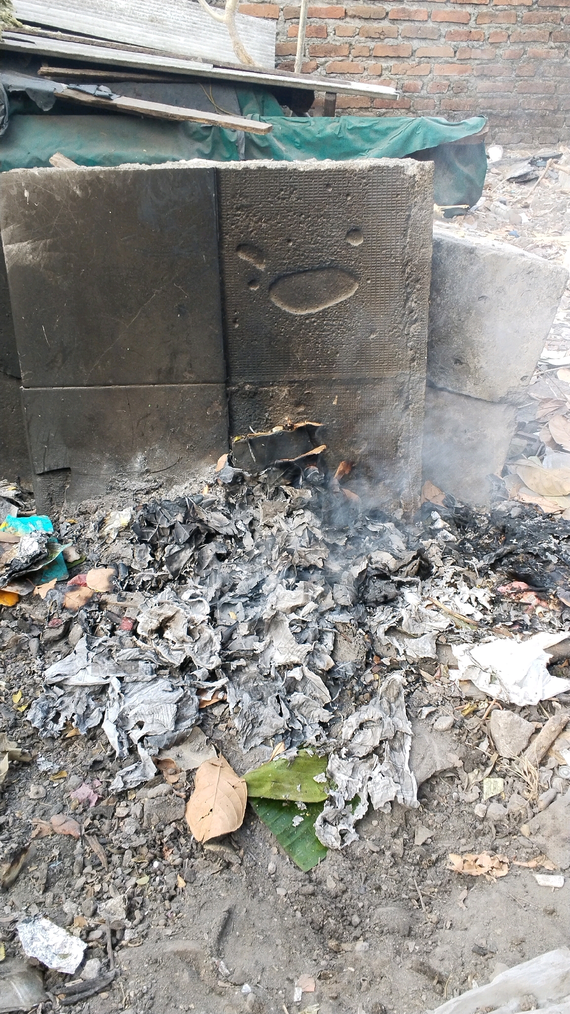 A pile of smoldering ashes and charred debris lies at the base of a large, dark, rectangular concrete structure. The concrete structure has a textured surface with what appear to be two indentations resembling eyes and a curved line beneath them, possibly a stylized face. To the right of this main structure, another large, rough-textured concrete block is visible. The ashes are spread across the ground, mixed with dirt, small stones, and a few scattered leaves, some of which are green and others brown and dry. Wisps of smoke rise from the smoldering material. In the background, more debris is visible, including what looks like corrugated metal sheeting and a brick wall. The scene suggests an outdoor area, possibly a construction site or a neglected urban space in Penompo, Indonesia, where waste is being burned. There are no people or animals visible in the image.