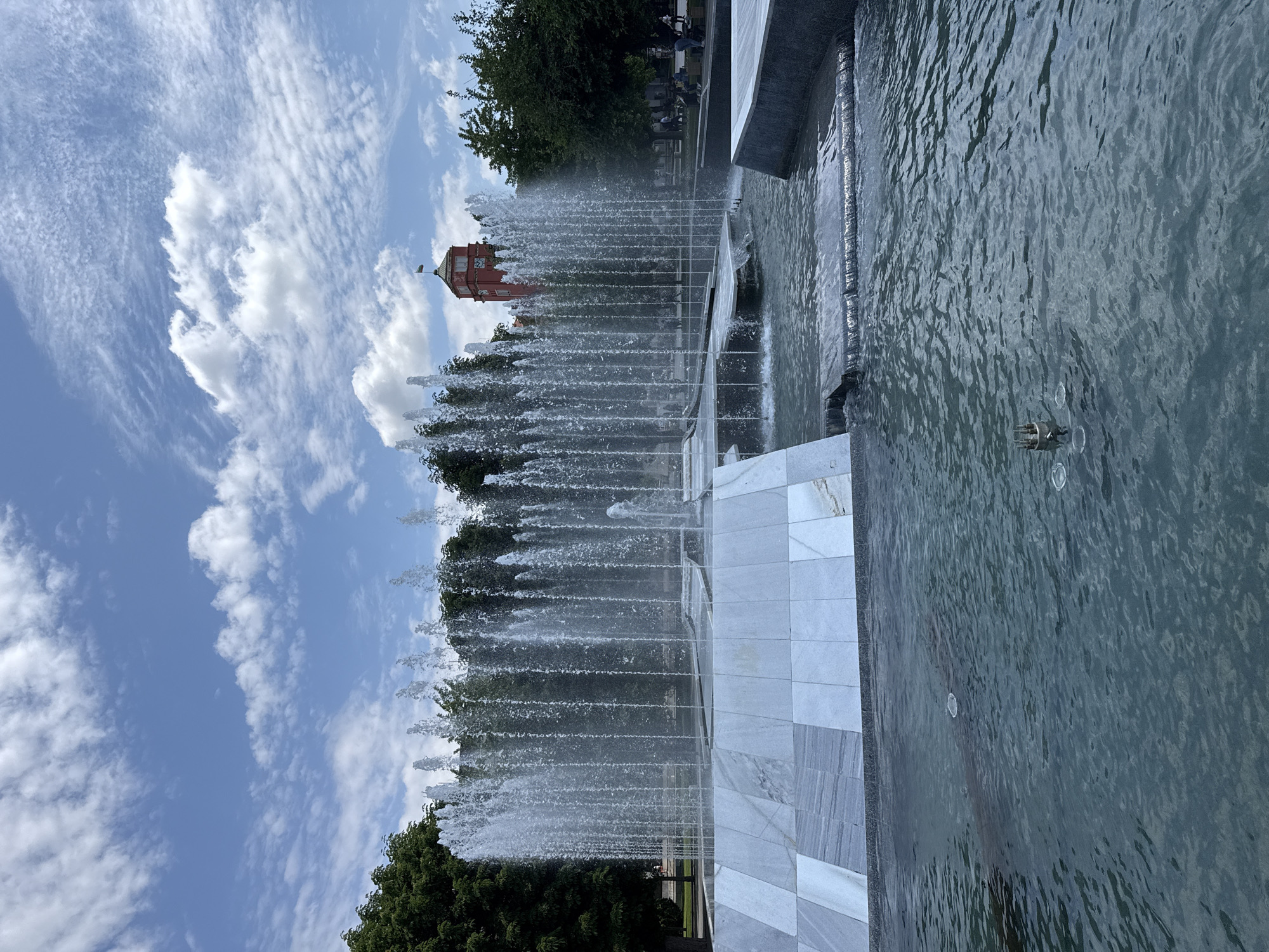 The image shows a water fountain in Pleven, Bulgaria, against a backdrop of a blue sky with white clouds and a red building. The fountain features multiple jets of water spraying upwards, creating a vertical display. The fountain structure is constructed of marble or a similar light-colored stone. Water flows from the jets into a pool of water. Vegetation, including trees, can be seen behind the fountain. The red building, possibly a tower, is visible in the distance, situated above the trees and fountain jets.