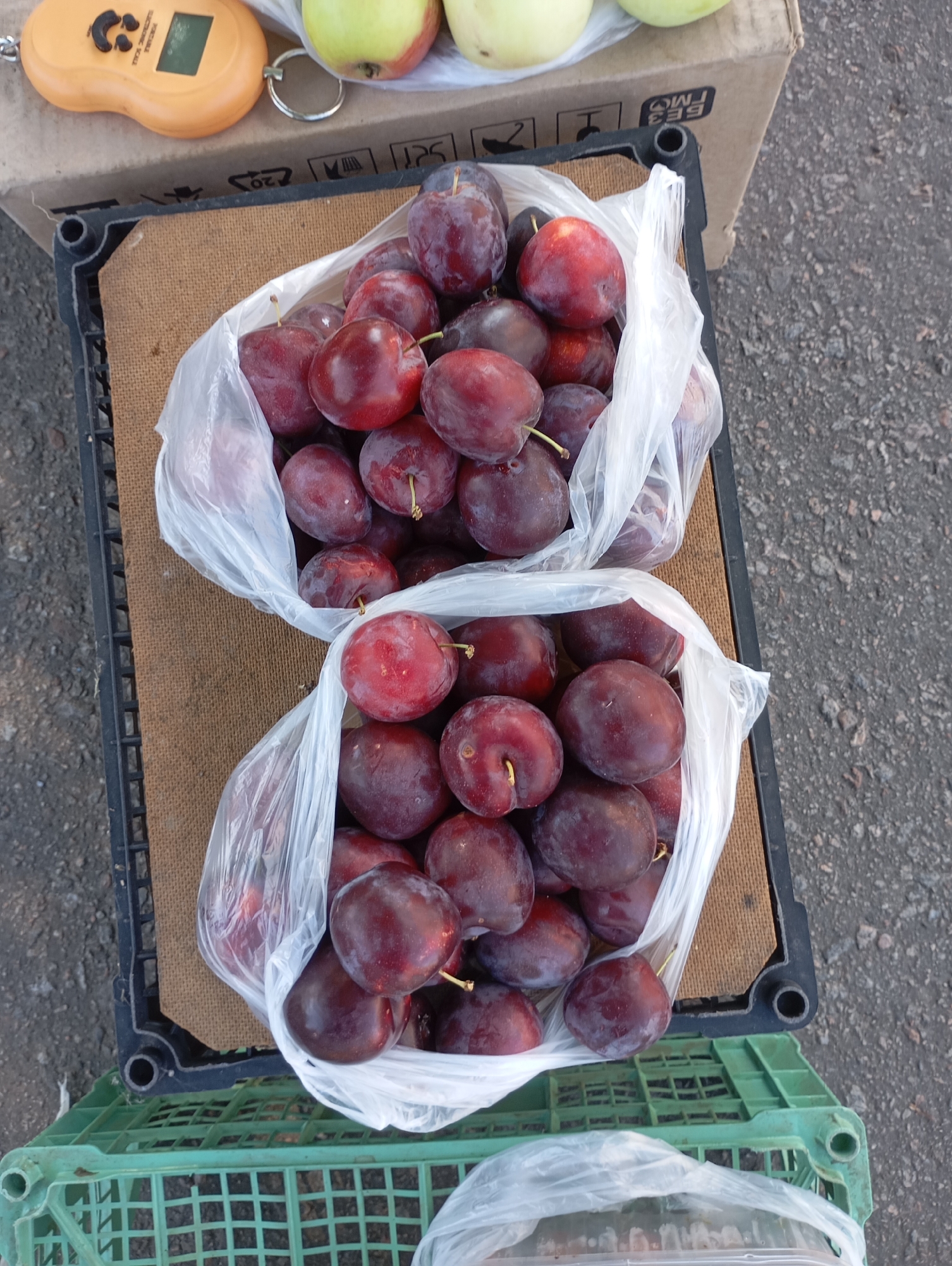 A high-angle shot shows a market stall with two plastic bags filled with plums. The bags are placed in a black crate on top of a cardboard box. In the background, there are some apples on the box and a portable digital scale. The ground appears to be asphalt. The scene is likely in the city of Horishni Plavni, Ukraine, given the context of the previous query.