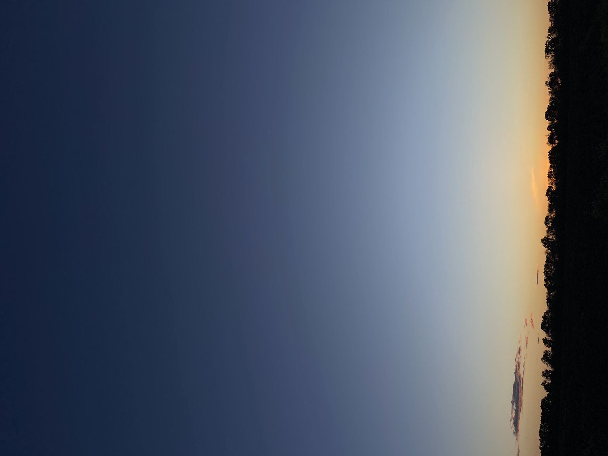The image depicts a horizontal view of the sky at dusk or dawn. The left side of the image shows a deep blue sky, transitioning to a lighter blue and eventually to a light orange near the right edge. The horizon line is defined by a dark silhouette of trees. The image does not provide sufficient information to confirm or deny that it was taken in the city of Popasne, Ukraine. No people, actions, or interactions are visible in the image.