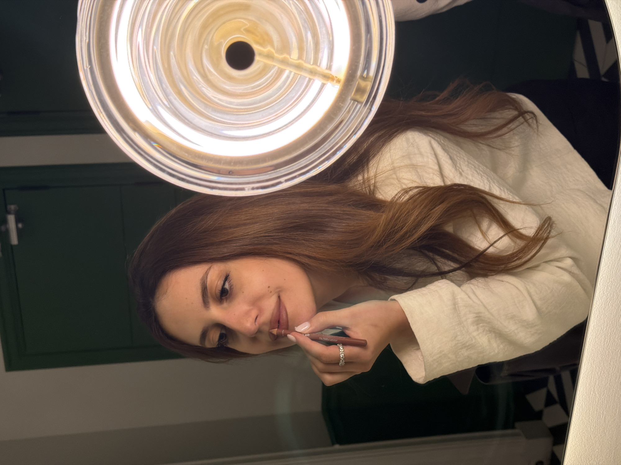 A person is reflected in a mirror while applying lip liner. The person has brown hair and is wearing a white top. The mirror reflects the underside of a circular light fixture. The background includes green cabinets and black-and-white tiled patterns. No identifying information indicates the location as Yerevan, Armenia.