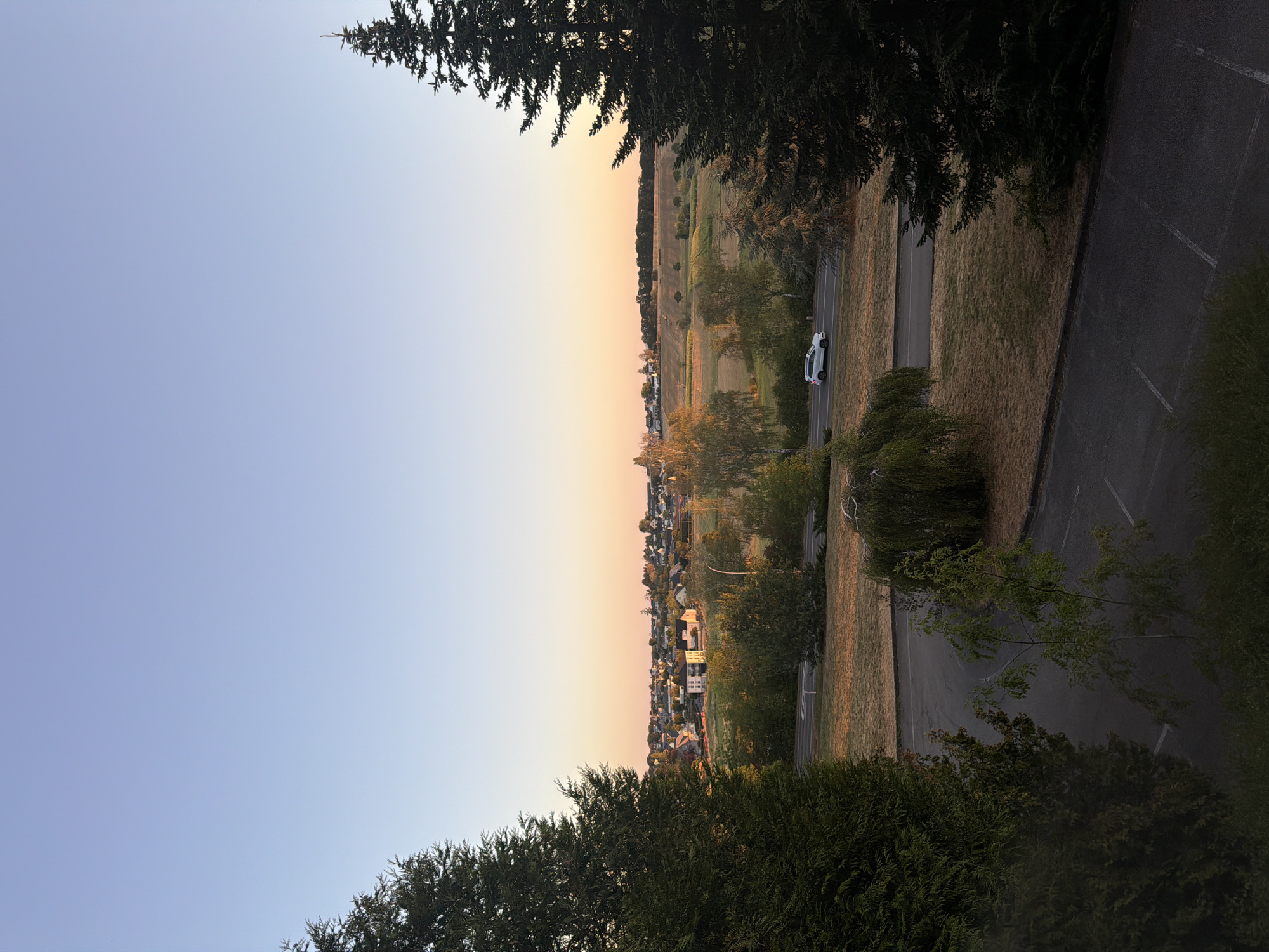 The image depicts a serene rural landscape at sunrise or sunset, likely in or near Leudelange, Luxembourg. The sky is a gradient of clear blue transitioning to soft orange and pink hues near the horizon. In the middle ground, a small town with white-roofed buildings is nestled amongst rolling hills. The fields surrounding the town are a mix of golden-brown grass and green patches, suggesting agricultural land. A paved road runs horizontally through the scene, with a white car driving on it.
Framing the landscape are two large evergreen trees, one on the left and one on the right, their dark green foliage contrasting with the lighter tones of the distance. In the foreground, a dark asphalt parking lot with white markings is visible, partially shaded by trees. A weeping willow tree with green foliage adds a touch of softness to the foreground. The overall impression is one of tranquility and natural beauty, with a touch of human habitation integrated into the environment.