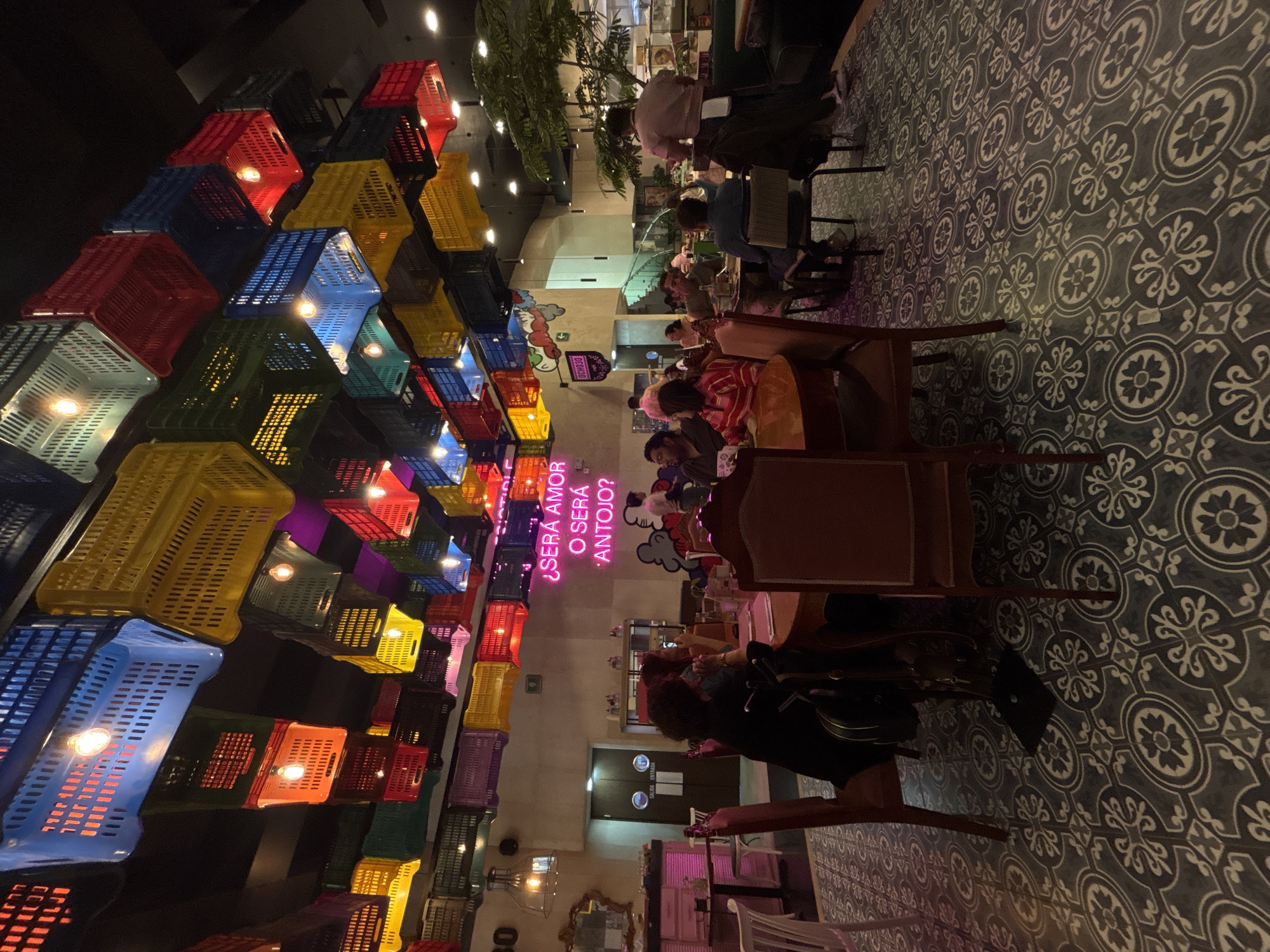 The image is taken inside a restaurant with overhead lighting. The walls are adorned with a column of colorful plastic crates, each containing a light. A pink neon sign, reading "Será amor o será antojo" in Spanish, is also mounted on the wall.
The restaurant appears to be busy, with patrons seated at tables and a waitperson serving. The floors are patterned with dark grey and white tiles. The image suggests an indoor location, possibly Puebla, Mexico based on the Spanish neon sign. The atmosphere appears lively and inviting.