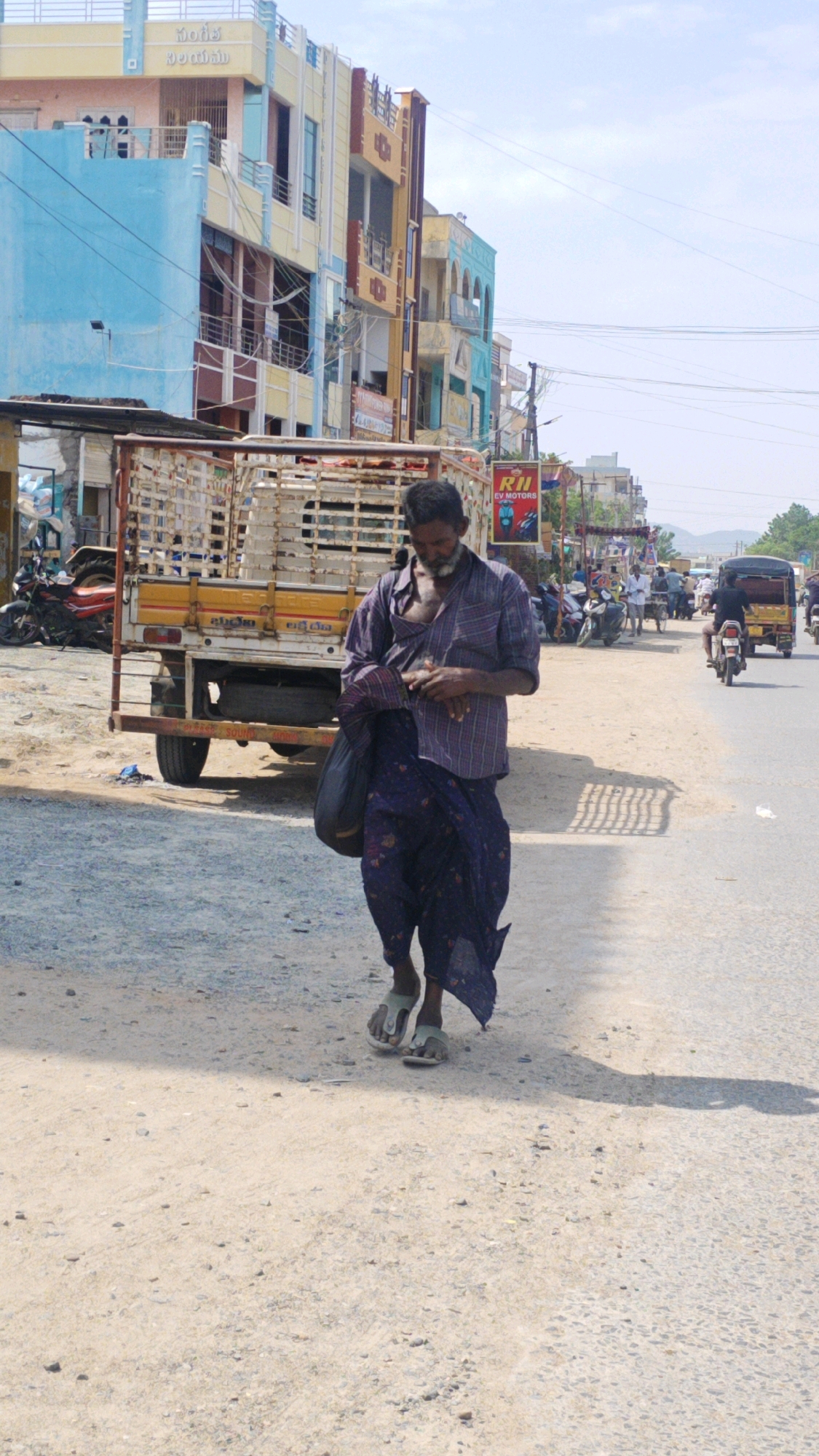 The image depicts a man walking on a dusty road in Pamur, India. He is wearing a purple checkered shirt, unbuttoned at the top, and a dark blue patterned lungi. A dark bag is slung over his left shoulder. He is wearing light-colored sandals. Behind him, a white truck with a caged bed is parked on the left side of the road. The truck has a yellow stripe along its side with writing. In the background, buildings with colorful facades line the right side of the road, appearing to be shops or residential units. A sign for "RII EV MOTORS" is visible on one of the buildings. Several motorcycles and an auto-rickshaw are also present on the road, with other pedestrians in the distance. The ground is covered in gravel and sand, and a shadow of the man and the truck is cast on the road. The sky is bright blue with some clouds.