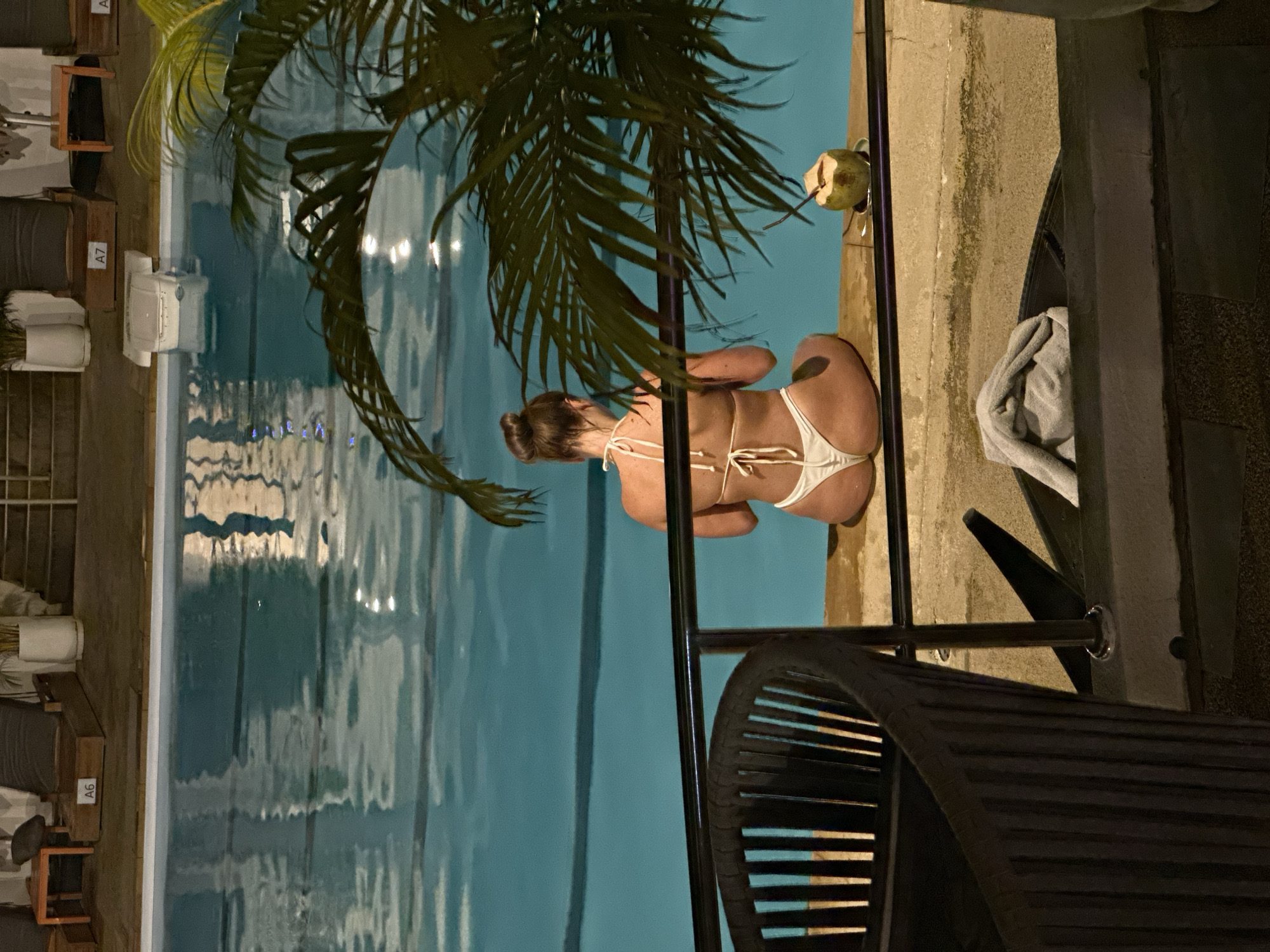 A woman in a white bikini is sitting on the edge of a swimming pool. She is facing away from the camera and has her hair in a bun. There is a coconut drink next to her on the pool edge. In the foreground, there is a black chair and a railing. The swimming pool water is illuminated by artificial lights, creating reflections on the surface. In the background, there are lounge chairs and potted plants. The scene appears to be at a resort or hotel.
