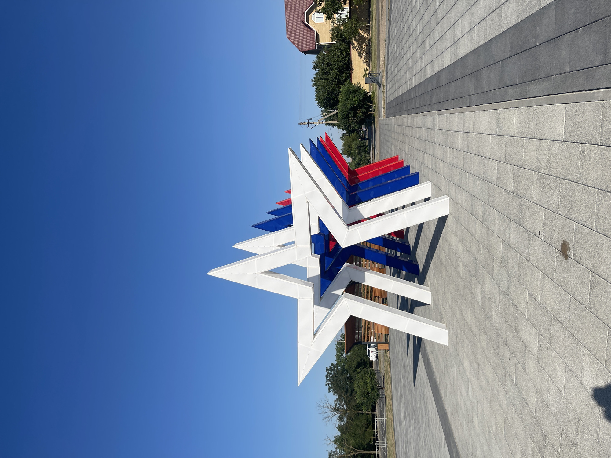 A row of five large, three-dimensional stars, painted white, blue, and red, stands in an outdoor public space. The stars are arranged in a line, receding into the background. The star in the foreground is white, followed by a blue star, then a red star, another blue star, and finally another red star. The stars are hollow and appear to be made of metal or a similar material. The ground in front of the stars is paved with large, light gray rectangular tiles. A darker gray paved strip runs parallel to the stars. In the background, there is a clear blue sky. To the left, there are trees and a white car parked near a building. To the right, there is a building with a red-tiled roof, more trees, and a utility pole. The image was taken in the city of Budyonnovsk, Russia.