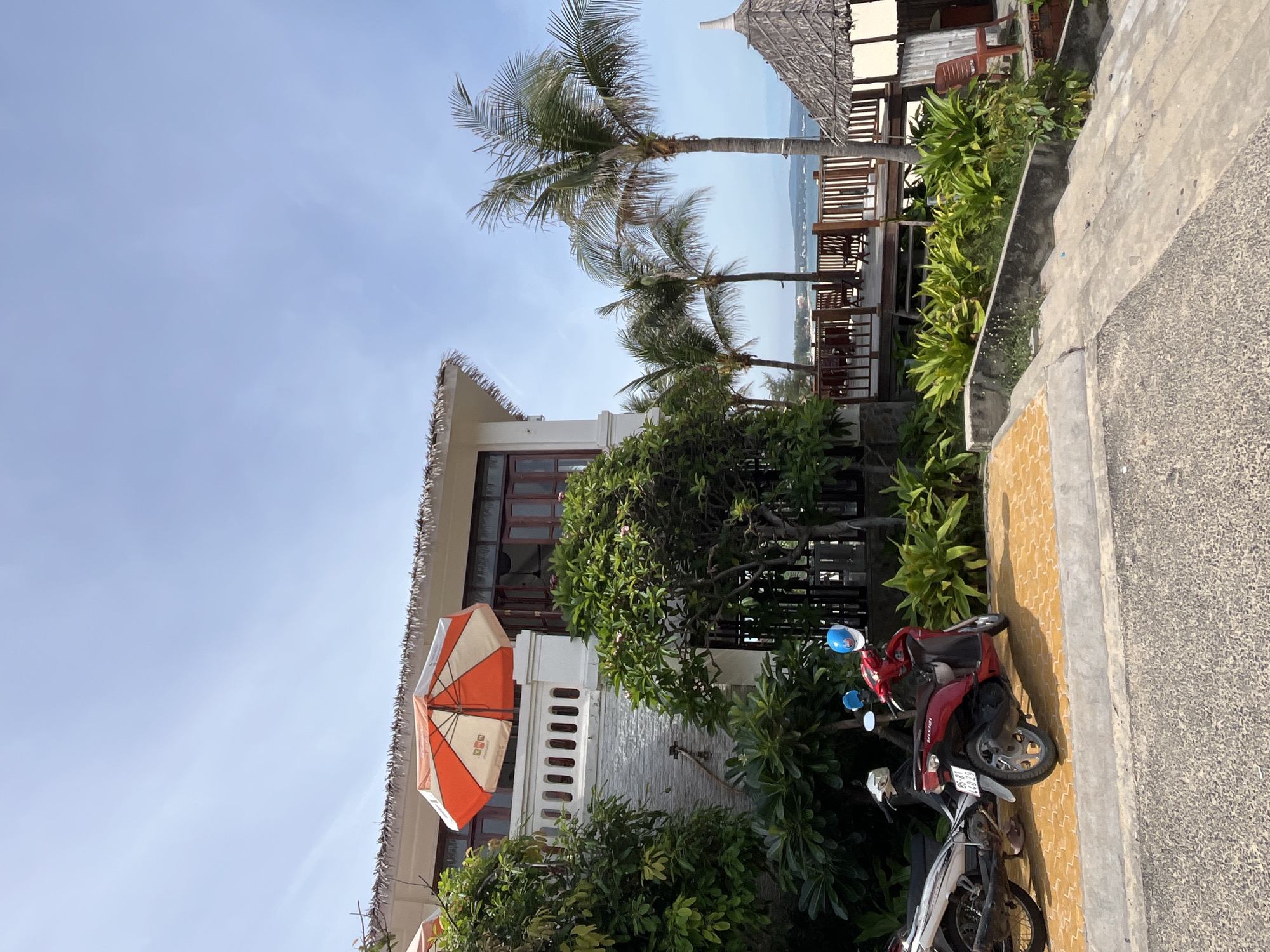 The image shows a tropical resort scene on a sunny day. In the foreground, there are two parked motorbikes on a yellow tiled surface. One is a red scooter with a blue helmet on the handlebars, and the other is a silver and white motorcycle. Behind the motorbikes, lush green foliage and trees surround a two-story building with a thatched roof overhang. The building has white walls, dark-paned windows, and a balcony with white railings. An orange and white umbrella is open on the balcony. Further back, a wooden deck with chairs and railings overlooks a body of water with a distant shoreline. Palm trees are scattered throughout the scene, and another thatched-roof structure is visible on the right side. The sky is clear and blue with a few wispy clouds. The location is identified as Phu Thuy Ward, Vietnam.