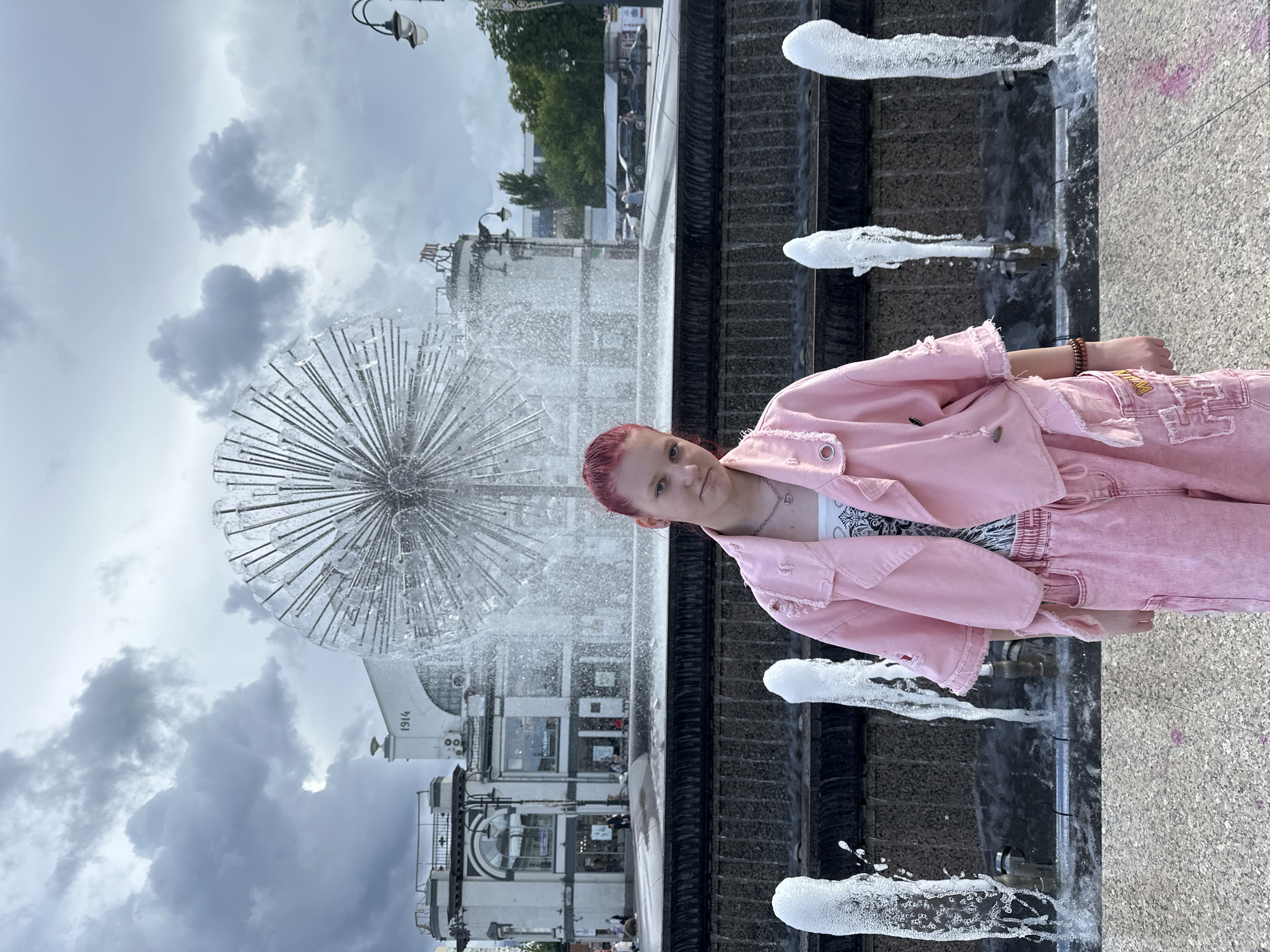 A person with reddish-pink hair stands next to a fountain in Saratov, Russia. The person is wearing a pink jacket and pants and is facing the camera. The fountain has a metal dandelion-shaped structure in the center with water spraying from it. The fountain's edge has multiple spouts shooting water upwards. Behind the fountain is a building with a sign that says "1914." The sky is overcast. The ground appears to be made of speckled concrete.