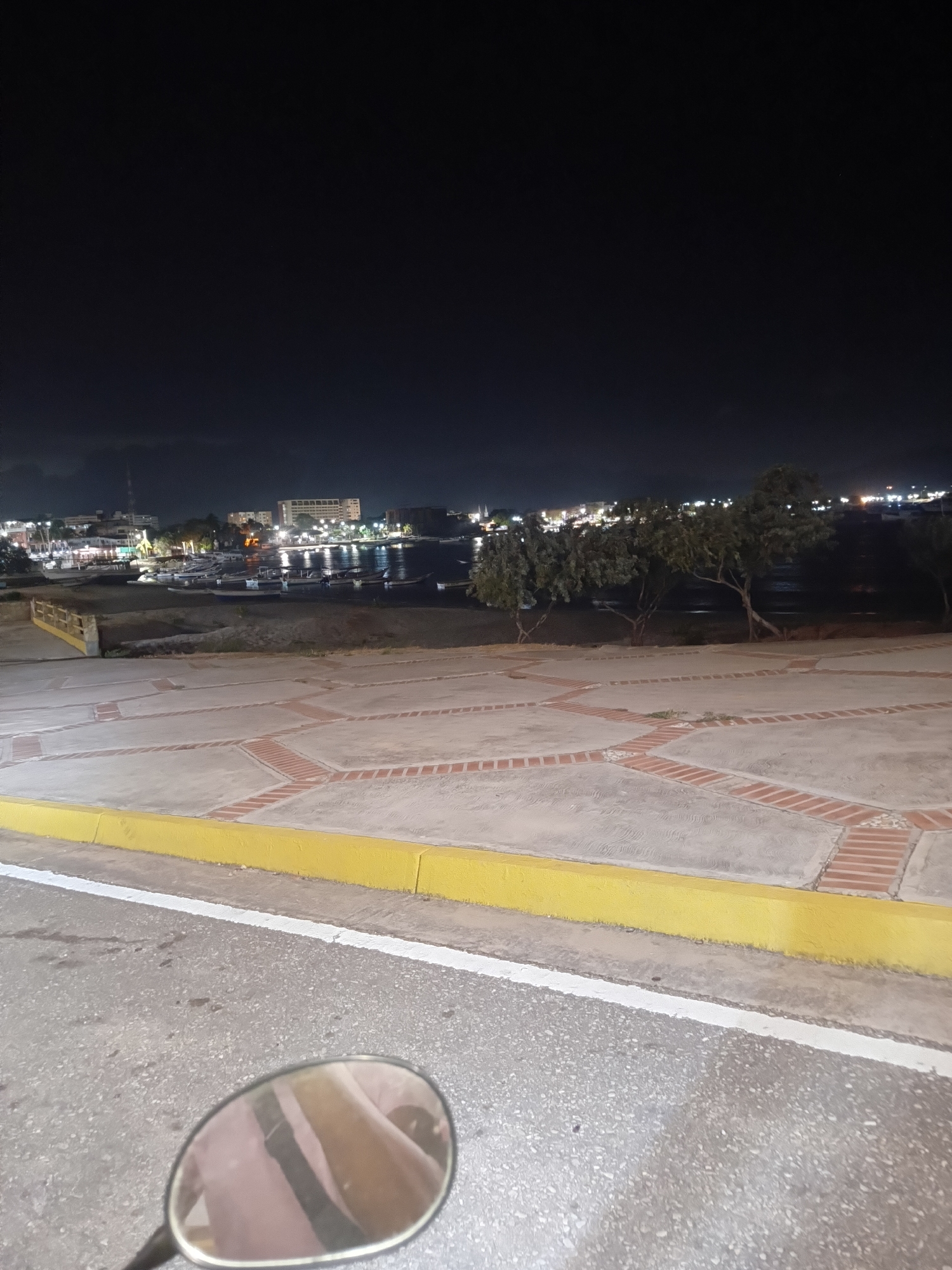 The image shows a nighttime view in Juan Griego, Venezuela. A paved area with brick patterns is in the foreground. In the distance, there is a harbor with boats and buildings lit up along the waterfront. The sky is dark. The scene is captured from a low angle, likely from a motorcycle, with the side mirror visible in the bottom center of the frame. There are trees located near the harbor. There are no people directly visible in the image.