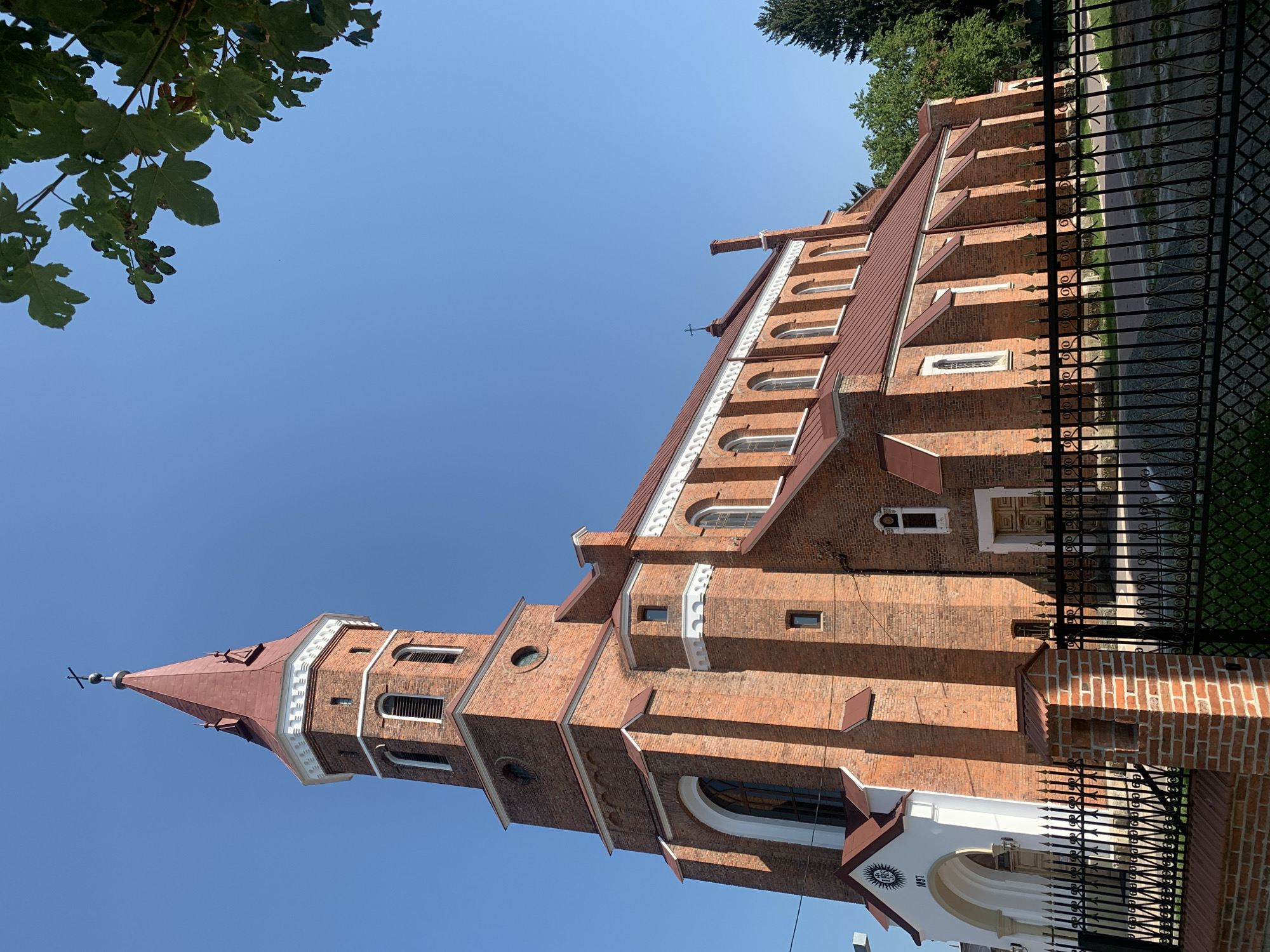 The image shows a red brick church with a red roof and a tall bell tower topped with a cross. The church has arched windows and a white facade on one side with the year "1897" inscribed above an arched entryway. A black metal fence runs along the front of the church, with a brick pillar in the foreground. The sky is clear and blue, and there are some green trees visible in the background. The church is likely located in Kolomyia, Ukraine, as indicated by the style and architectural elements often seen in Ukrainian churches.