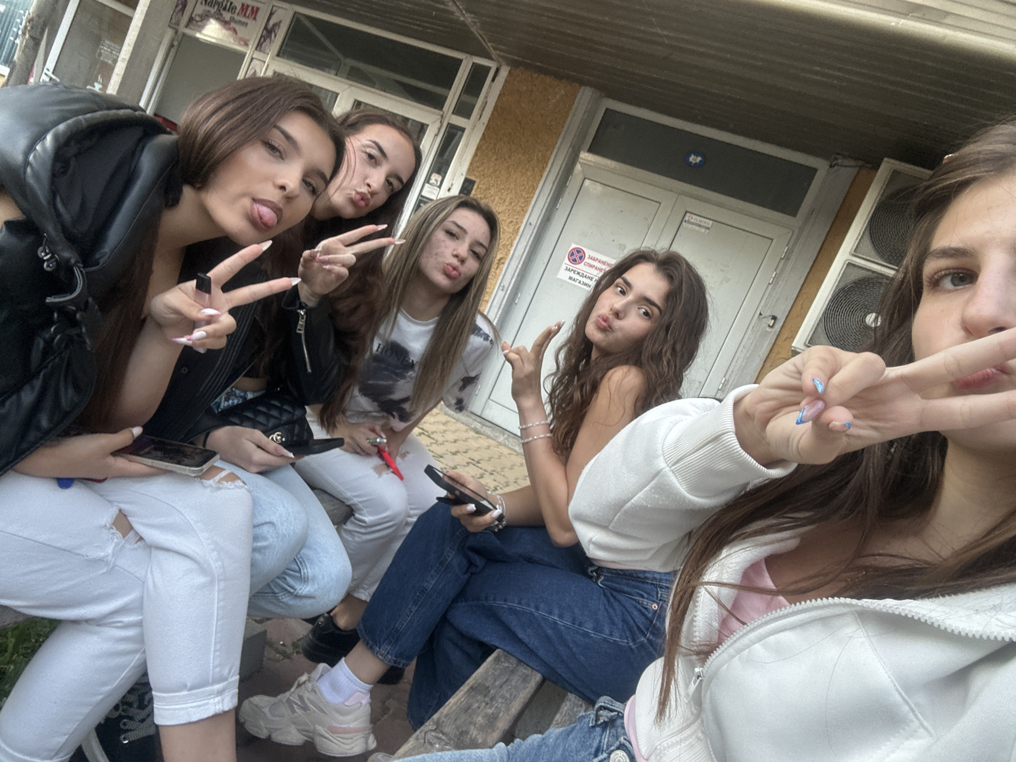 In Shumen, Bulgaria, a group of five young women are gathered outdoors, possibly near a commercial building with a textured facade and large double doors. The women are dressed casually in jeans and tops, some wearing jackets. Three of the women are posing for the camera, sticking out their tongues, making peace signs, and puckering their lips. Two of the women are holding smartphones, and one appears to be recording or taking photos. The environment suggests a public space, possibly an urban area, with a wooden bench visible beneath them. The overall atmosphere is informal and social.