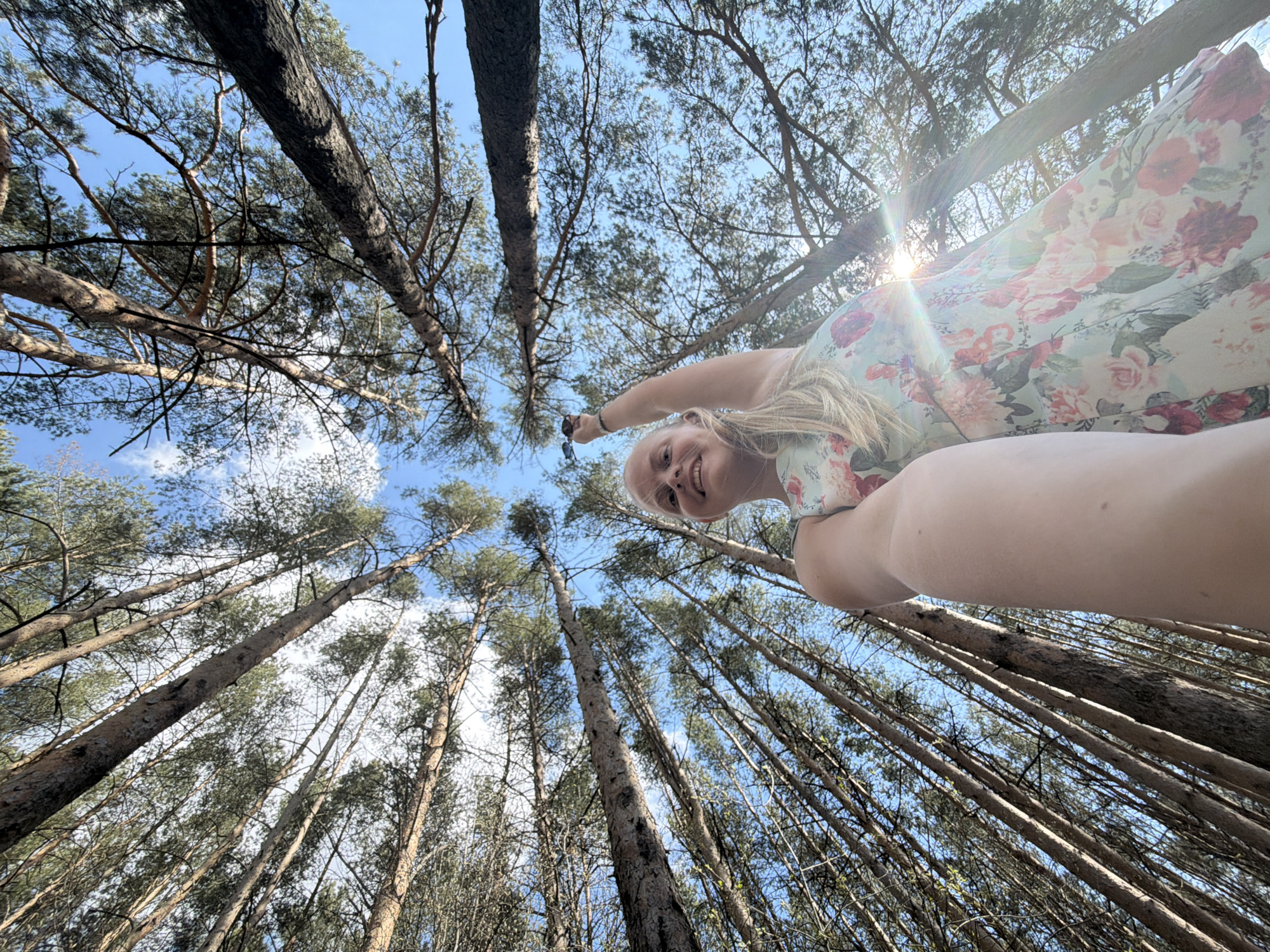 A wide-angle, upward-facing photograph depicts a fair-skinned woman in a floral dress amidst a forest of tall, slender trees, possibly in Izhevsk, Russia. The trees, with their textured bark, reach toward a partially visible blue sky. Sunlight peeks through the canopy. The woman is in the foreground, her face smiling towards the camera, holding a pair of sunglasses. The perspective is from below, looking up at the woman and the trees, accentuating their height.