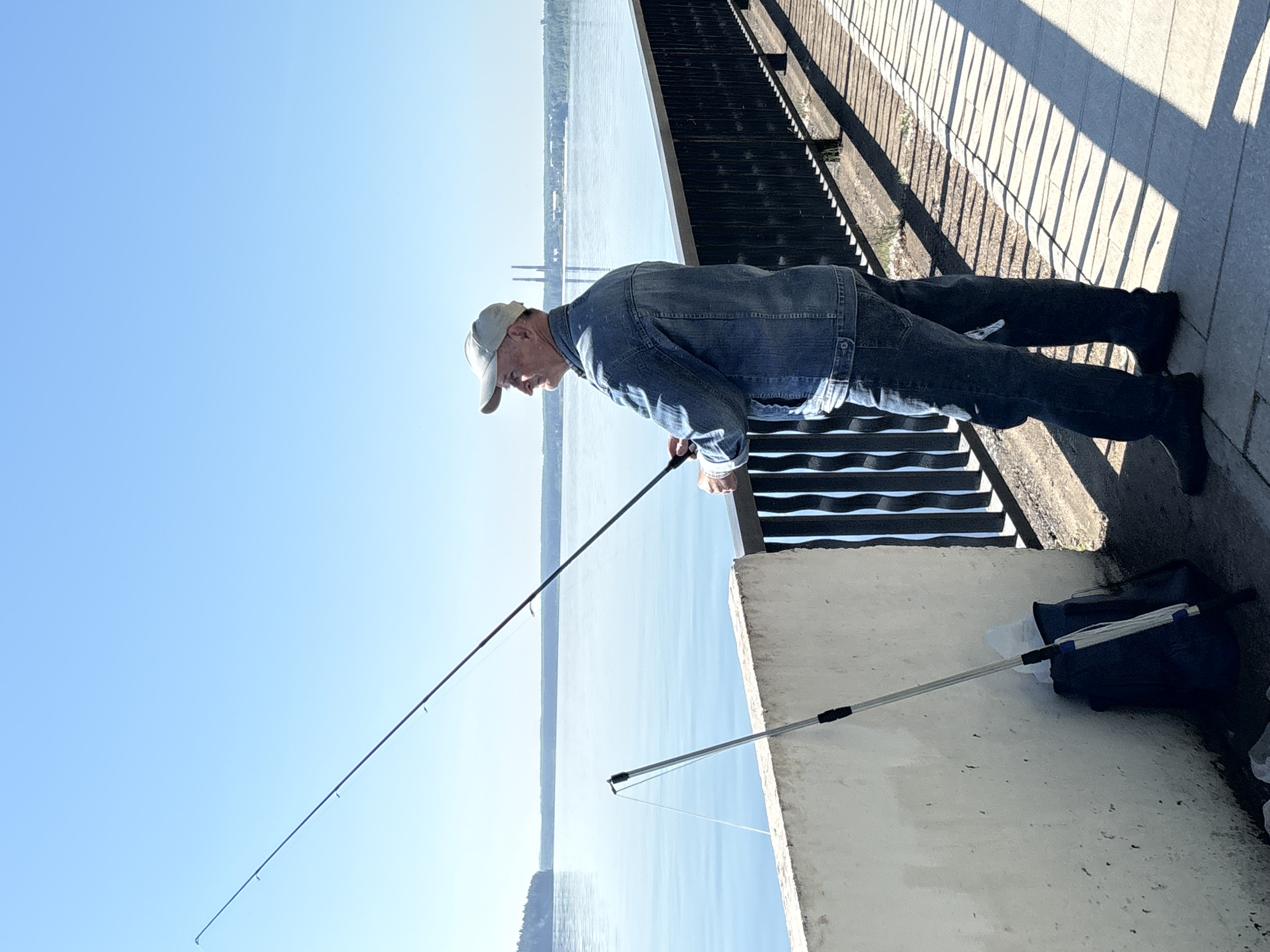 A man, appearing to be of Russian ethnicity and in his late 60s, is fishing on a sunny day along the embankment of the Kama River in Chaikovsky, Russia. He is wearing a denim jacket, jeans, and a light-colored baseball cap. The man is holding a fishing rod, with the line cast into the water. Next to him, leaning against a white concrete barrier, is another fishing rod, a dark blue backpack, and a white plastic bag. The embankment is paved with light-colored tiles, and a black metal railing runs along the edge of the river. The river is calm and reflects the clear blue sky. In the distance, across the river, industrial structures and green treelines are visible. The scene suggests a peaceful recreational activity by the water.