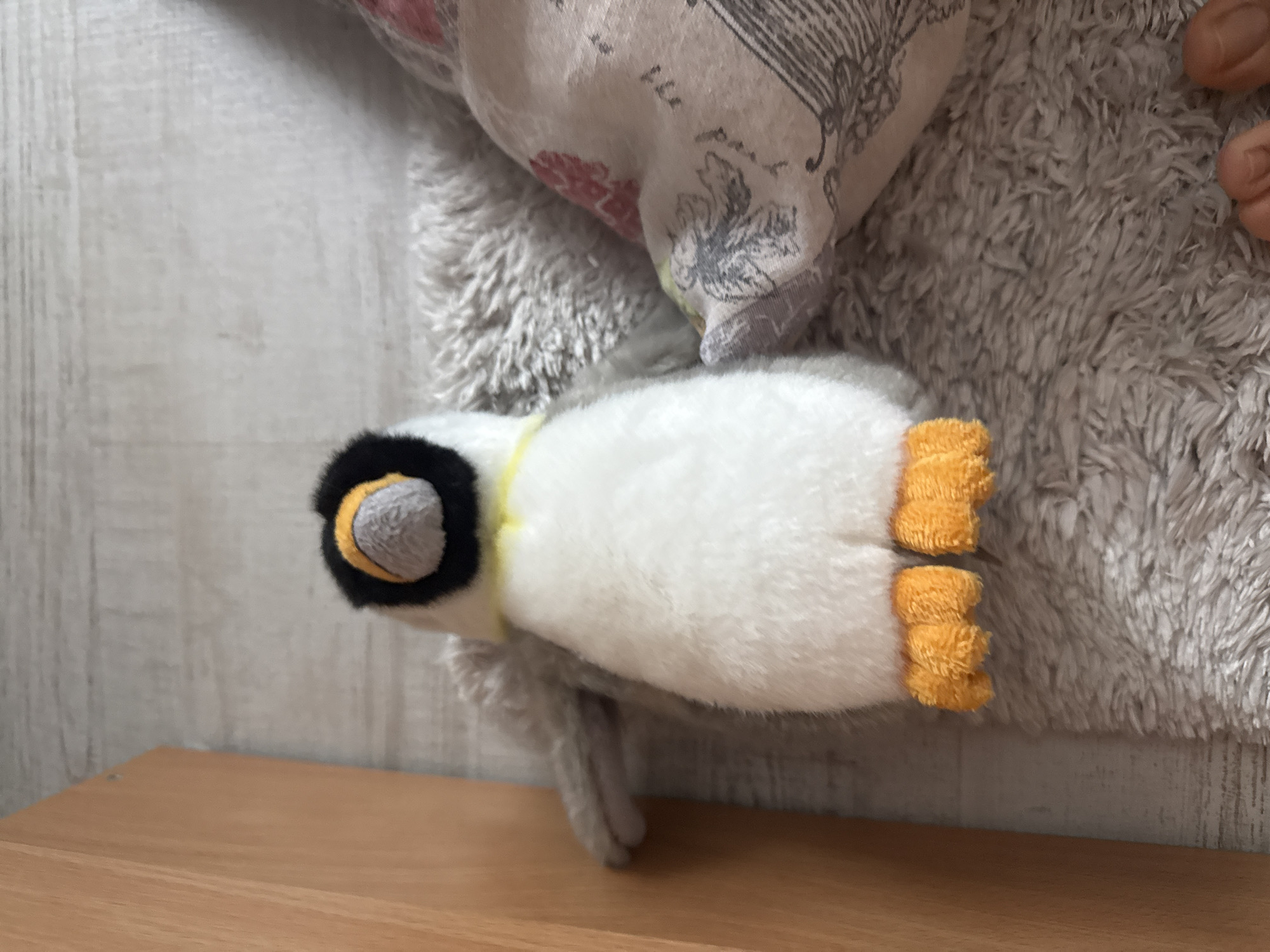 The image depicts a plush penguin toy, laying on a wooden surface, likely a table or shelf. The penguin is white with a black head and yellow accents around the eyes and on its feet. It appears to be nestled among soft, fuzzy fabrics. The specific location in Blagoevgrad, Bulgaria is not identifiable from the image.