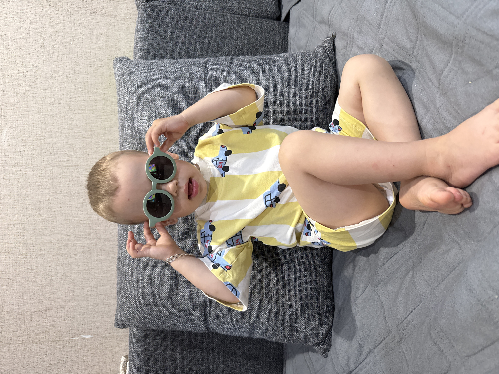 A young child lies on a gray couch and a gray pillow, wearing yellow and white striped pajamas with a car pattern and green sunglasses, partially touching the sunglasses with their right hand. The photo was taken indoors, and the location may be Rusanovo, Russia.