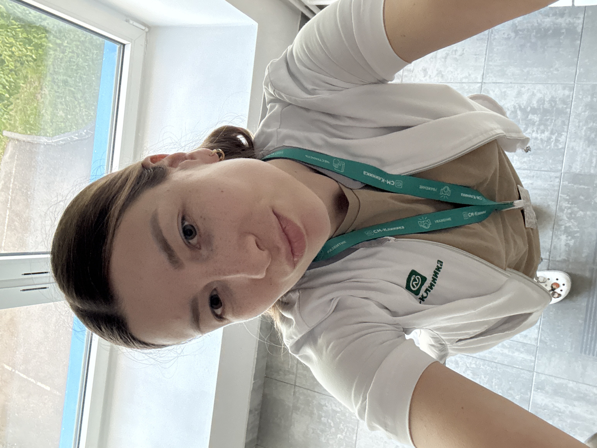 A young woman takes a high-angle selfie, looking directly at the camera with a neutral expression. Her dark brown hair is pulled back. She is wearing a light-colored, likely white, zip-up jacket over a tan top. Around her neck is a teal lanyard prominently displaying text in Cyrillic: "СМ-Клиника" (SM-Clinic), "УВАЖЕНИЕ" (RESPECT), "ЧЕСТНОСТЬ" (HONESTY), and "РАЗВИТИЕ" (DEVELOPMENT). The same "СМ-Клиника" branding and a logo are also visible on the front of her jacket. On her right foot, a white Croc-style shoe adorned with small charms is visible.
The scene is an indoor setting, likely a hallway or corridor within a building. To the left, a window provides a glimpse of the outdoors, showing green grass and what appears to be a paved path under bright daylight, indicating it is daytime. The floor is tiled with large grey squares, and the walls are white. The consistent branding "SM-Clinic" on both her lanyard and jacket strongly suggests she is at a medical facility or clinic, located in Moscow, Russia.