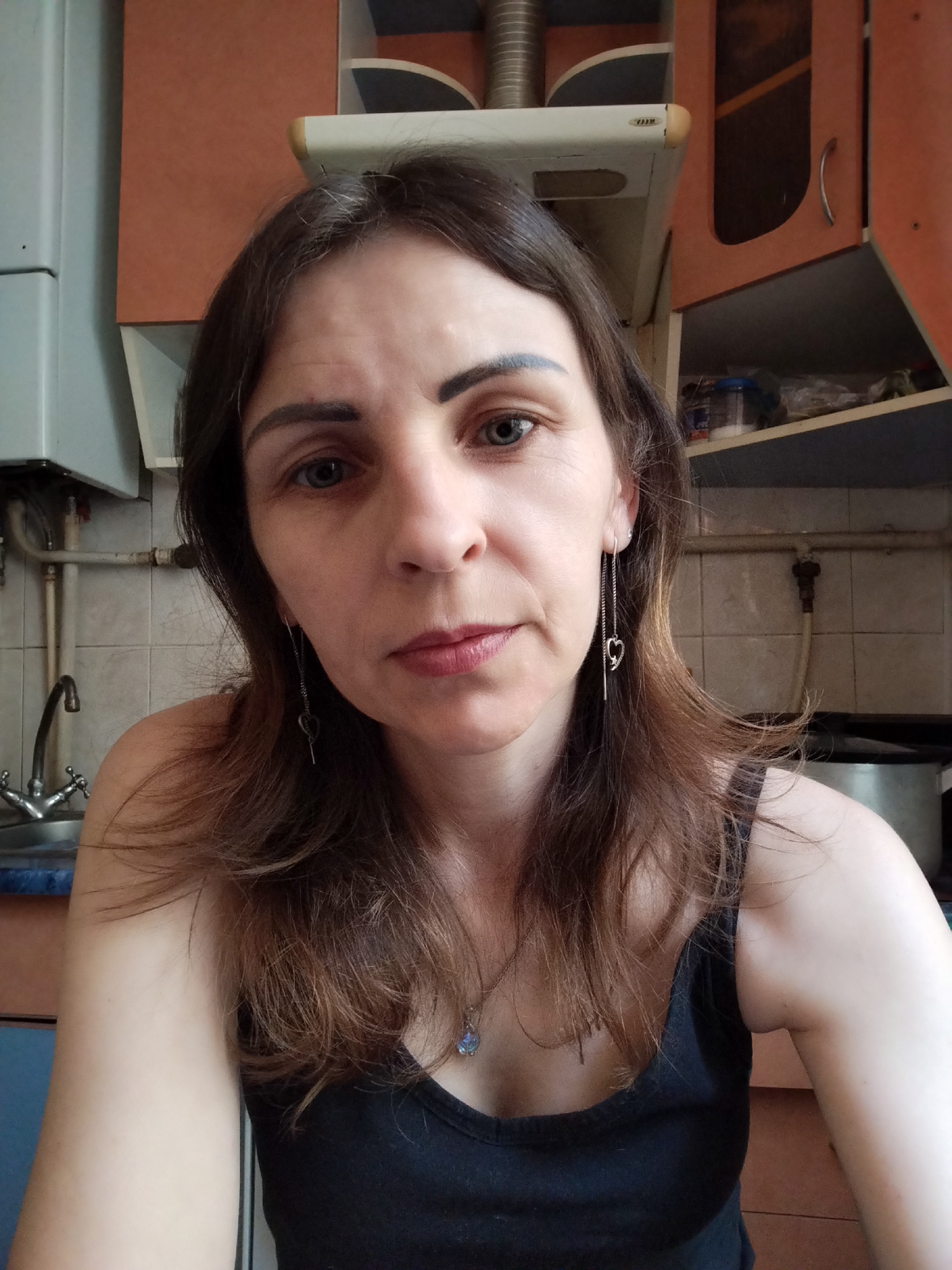 This is a selfie taken by a woman in what appears to be a kitchen. The woman has brown hair and is wearing a black tank top. She is wearing dangling earrings with heart-shaped charms. In the background, there are kitchen cabinets, a sink with a faucet, and pipes. The lighting suggests it is daytime. The specific location in Vinnytsia, Ukraine, is not determinable from the image alone.