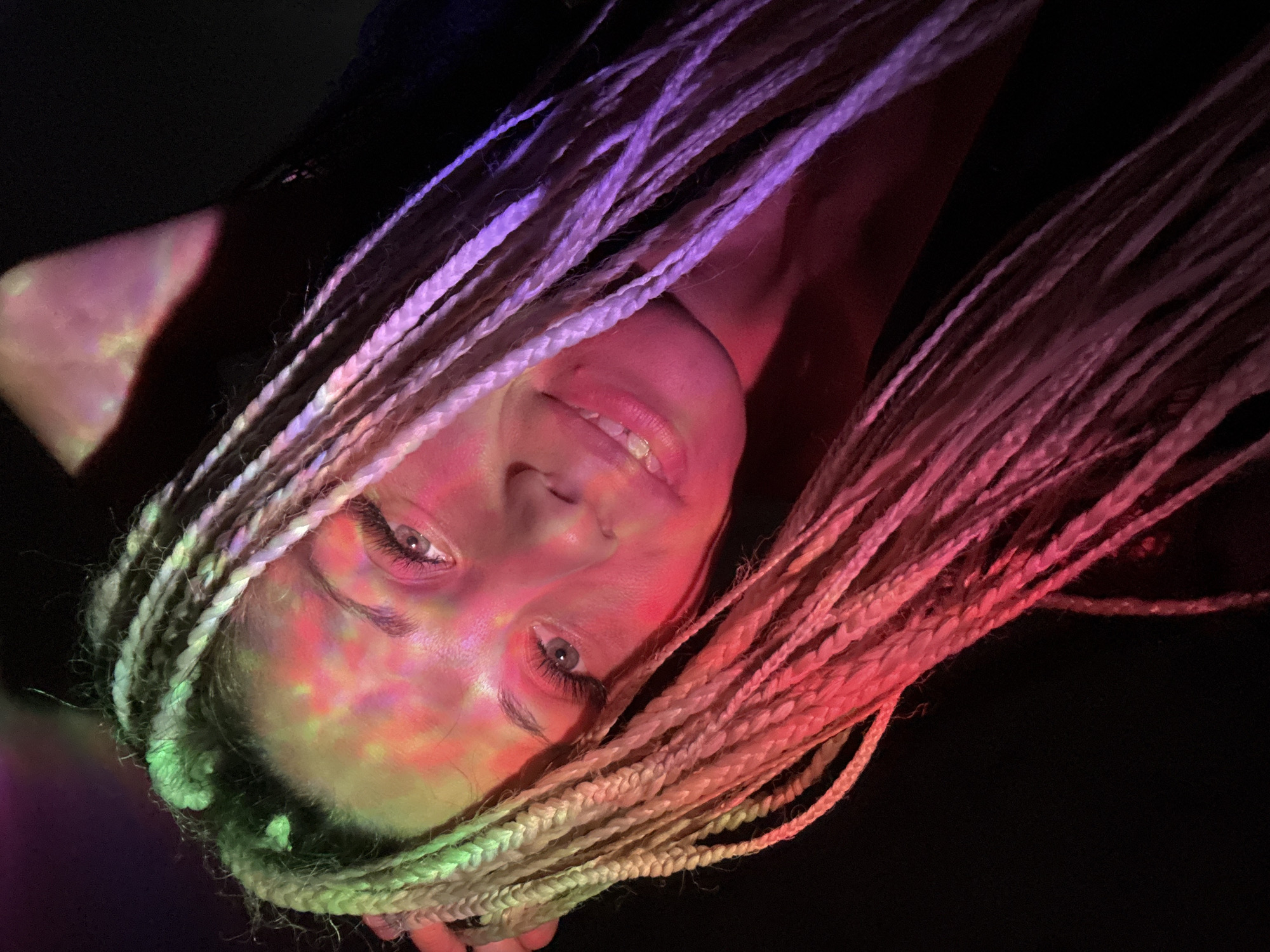 The image is a close-up, low-angle selfie of a young Caucasian woman with blonde dreadlocks. She is positioned in the lower right quadrant of the frame, looking towards the camera with a slight smile. Her face is illuminated by colorful, abstract light projections, with hues of pink, red, and green prominently visible on her forehead, cheek, and hair. Her eyes are open and her makeup, including prominent false eyelashes, is visible. She is wearing dark clothing. The background is predominantly dark, suggesting a dimly lit environment, possibly a club or a room with colored lighting effects. The image does not provide any information about the location in Zhukovo, Russia, or any specific context beyond the visual elements.