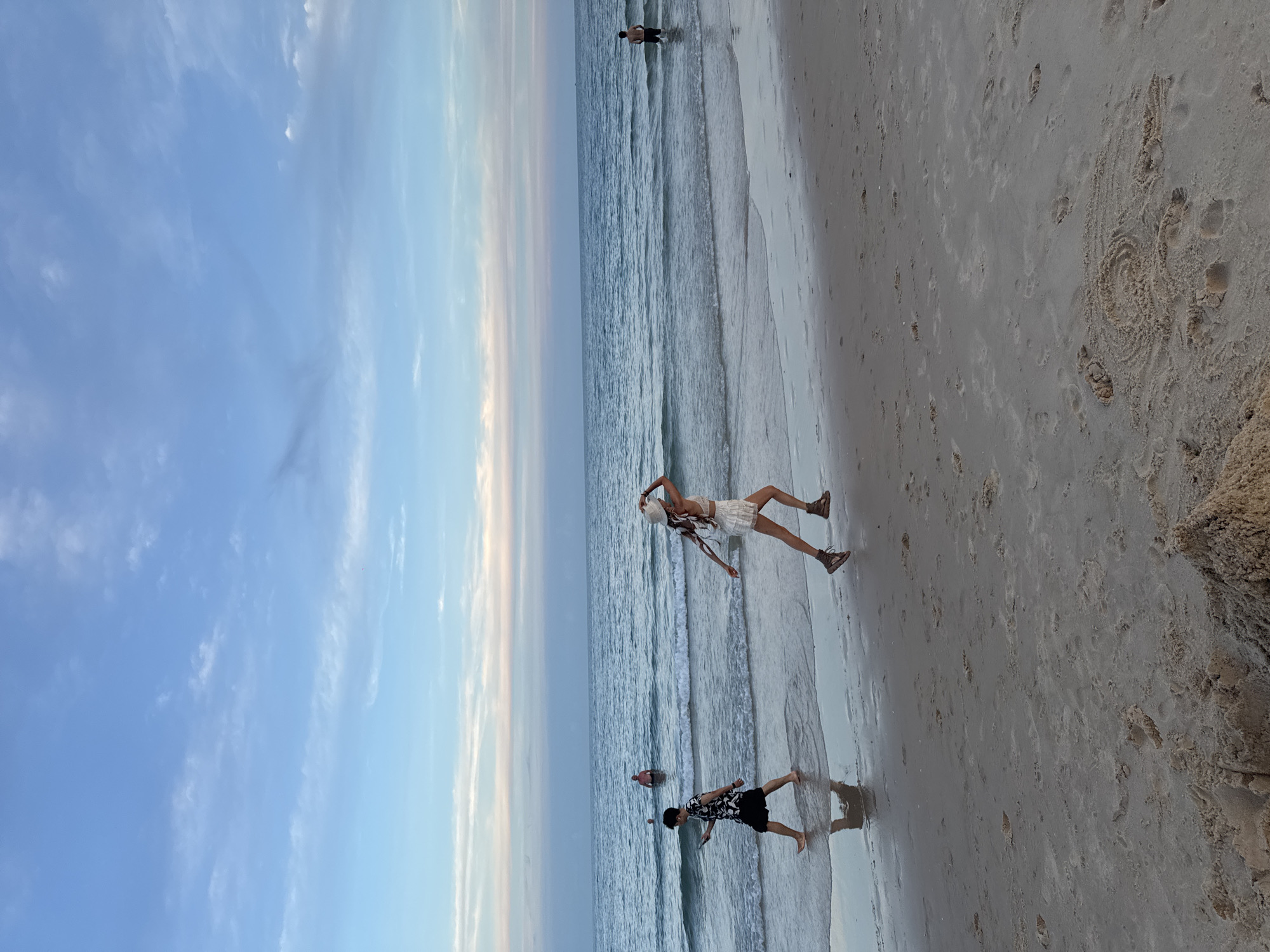 This image depicts a beach scene at dusk in Phu Thuy Ward, Vietnam. The sky is a gradient of pale blue to soft pink and orange hues, indicating the sun is setting. The ocean is calm with gentle waves lapping at the shore. In the foreground, a sandy beach is visible with footprints and some debris.
Several people are present. On the left, a young man with dark hair is walking along the water's edge, looking at his phone. He is wearing a black and white patterned short-sleeved shirt and black shorts. His reflection is visible in the shallow water.
To his right, a person, appearing to be a woman, is running along the beach. She is wearing a white hat, a white top, and white ruffled shorts, along with brown shoes. She is holding her right hand up to her hat.
Further out in the water, three individuals can be seen. One person is standing in the water up to their chest, facing away from the camera. Two other individuals are partially submerged in the water near the shore.
The overall atmosphere is serene and tranquil, capturing a moment of relaxation and activity on a Vietnamese beach as day transitions into night.