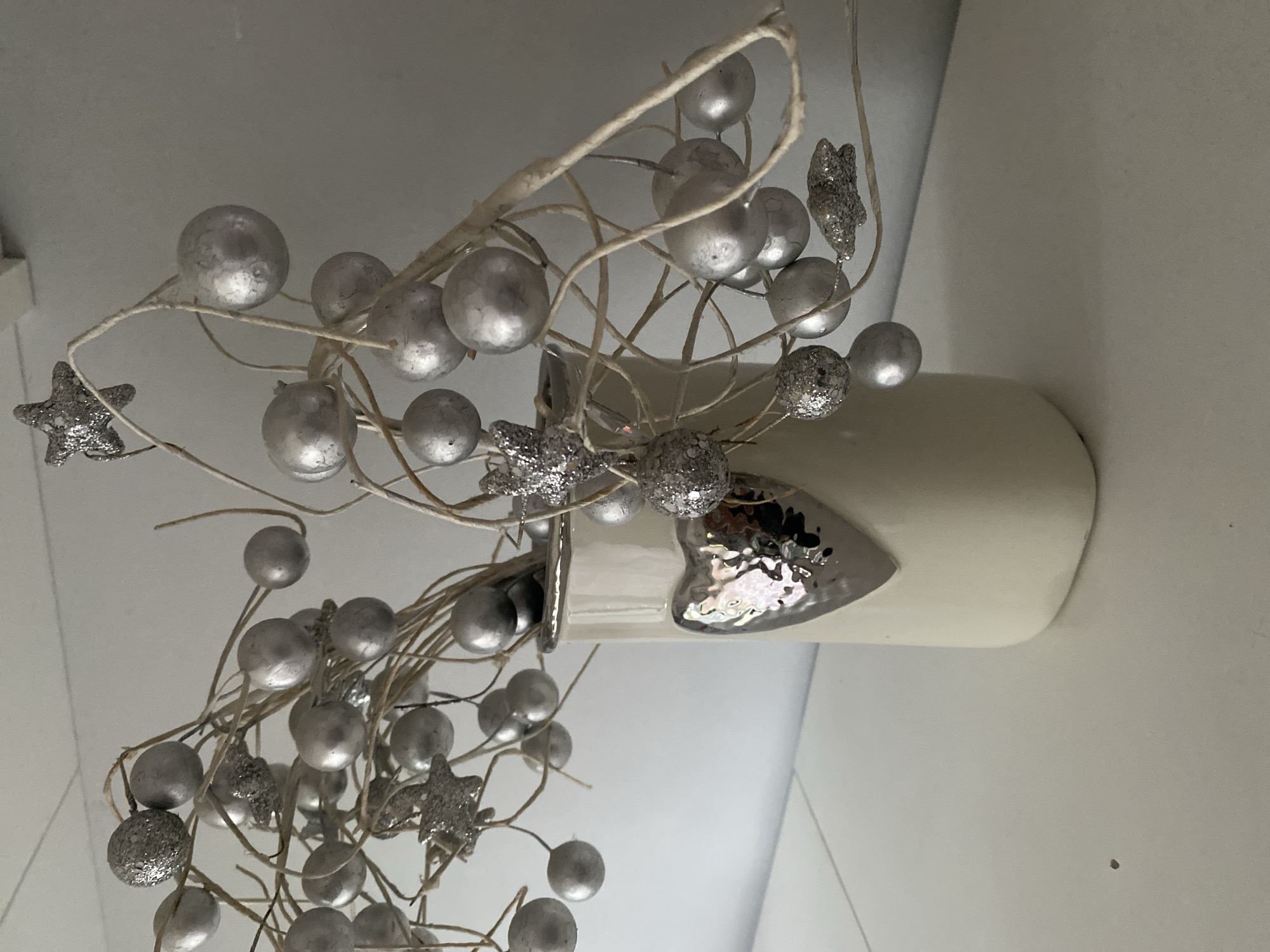 A close-up, eye-level shot shows a decorative arrangement of silver baubles and glittery stars on natural twigs, placed in a cream-colored ceramic vase. The vase has a textured silver heart embellishment on its front. The arrangement is positioned on a white surface, likely a shelf or table, against a neutral, light-colored background. The image is a still life, with no people or actions depicted. The lighting is soft and diffused, creating subtle shadows. The scene is indoors, in a clean, minimalist setting. The location in the city of Wrocław, Poland is not discernible from the provided image.
